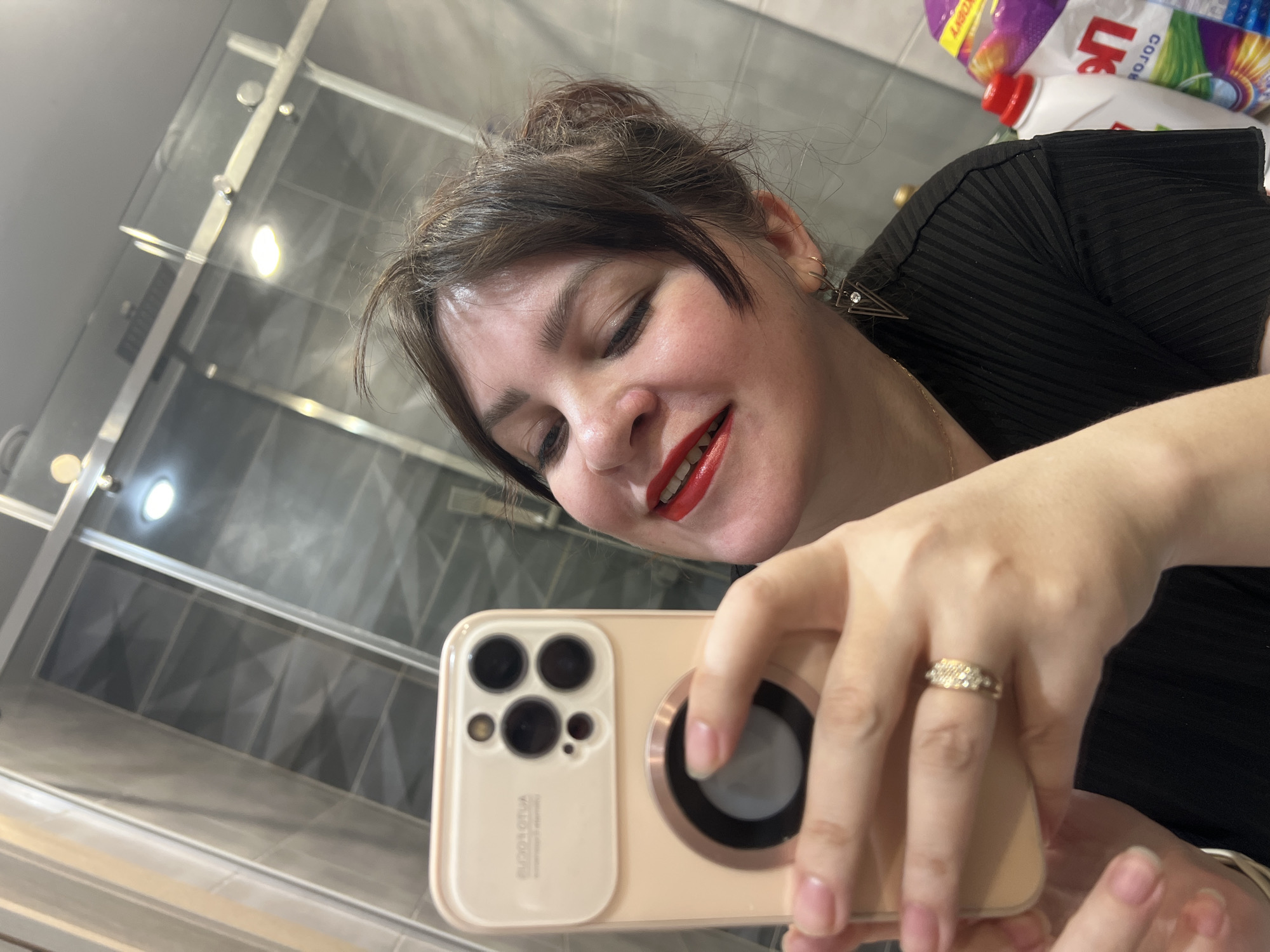 An adult female is depicted taking a mirror selfie in a modern bathroom. She is shown from the mid-chest up, with dark brown hair, red lipstick, and her eyes closed or partially closed as she smiles. She wears a black ribbed top, a gold-colored ring with multiple small stones on her right ring finger, and a dark geometric triangular earring with a small white stone in her right ear. Her left hand holds a light-colored smartphone, equipped with a multi-lens camera module and a circular accessory on its back; "DUOOZ" or similar text is partially visible on the camera cover. The bathroom features grey tiled walls with a textured, possibly geometric, pattern. A clear glass shower enclosure with metal hardware is reflected behind her. Additionally, reflections reveal overhead recessed lighting and, in the lower right, partially visible household items including a white bottle with a red cap and a purple and yellow package displaying Cyrillic text, consistent with a Russian-speaking region. The specific location in the city of Noviy, Russia, cannot be definitively confirmed from the visual evidence provided.