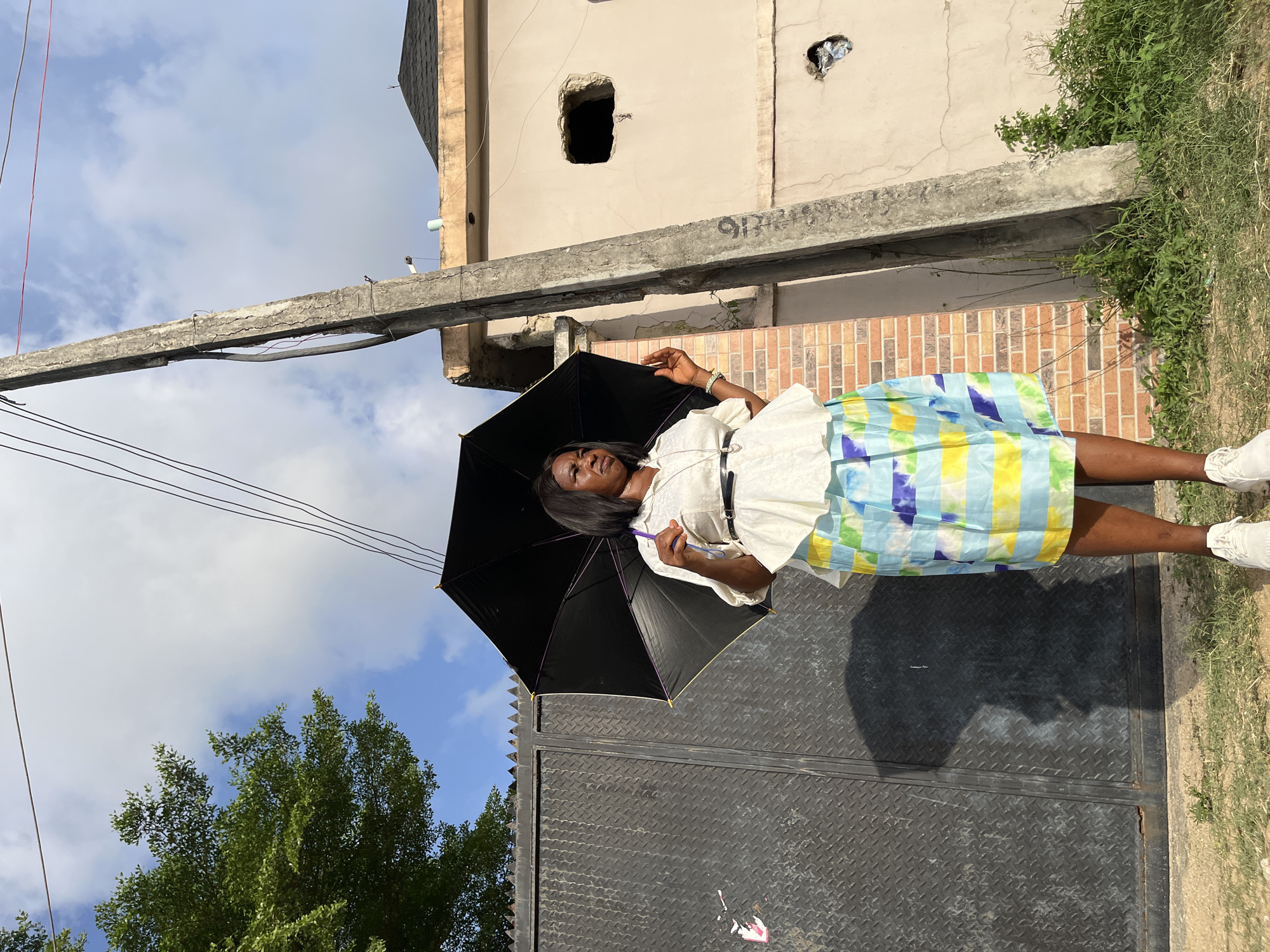 An adult female stands outdoors in Iranla, Nigeria, holding an open black umbrella overhead. She is positioned facing slightly right of the viewer, looking towards her left. She wears a white blouse with a black belt, a knee-length skirt patterned with light blue, yellow, and darker blue stripes, and white athletic shoes. Her shadow, including that of the umbrella, is cast clearly onto a large, dark gray diamond-plate metal gate to her immediate left. The gate features spiked anti-climb elements at its top. To her right stands a weathered concrete utility pole, exhibiting cracks and supporting multiple dark electrical wires overhead. A light-colored building with a dark, possibly tiled roof is visible in the background, to the right of the pole. This building features a large, irregular rectangular opening at mid-height and a smaller, similar opening lower down, containing reflective debris. A section of a brick wall is visible between the gate and the utility pole. The ground consists of sparse dry grass, dirt, and green weeds. A green deciduous tree is partially visible on the far left, under a partially cloudy blue sky.