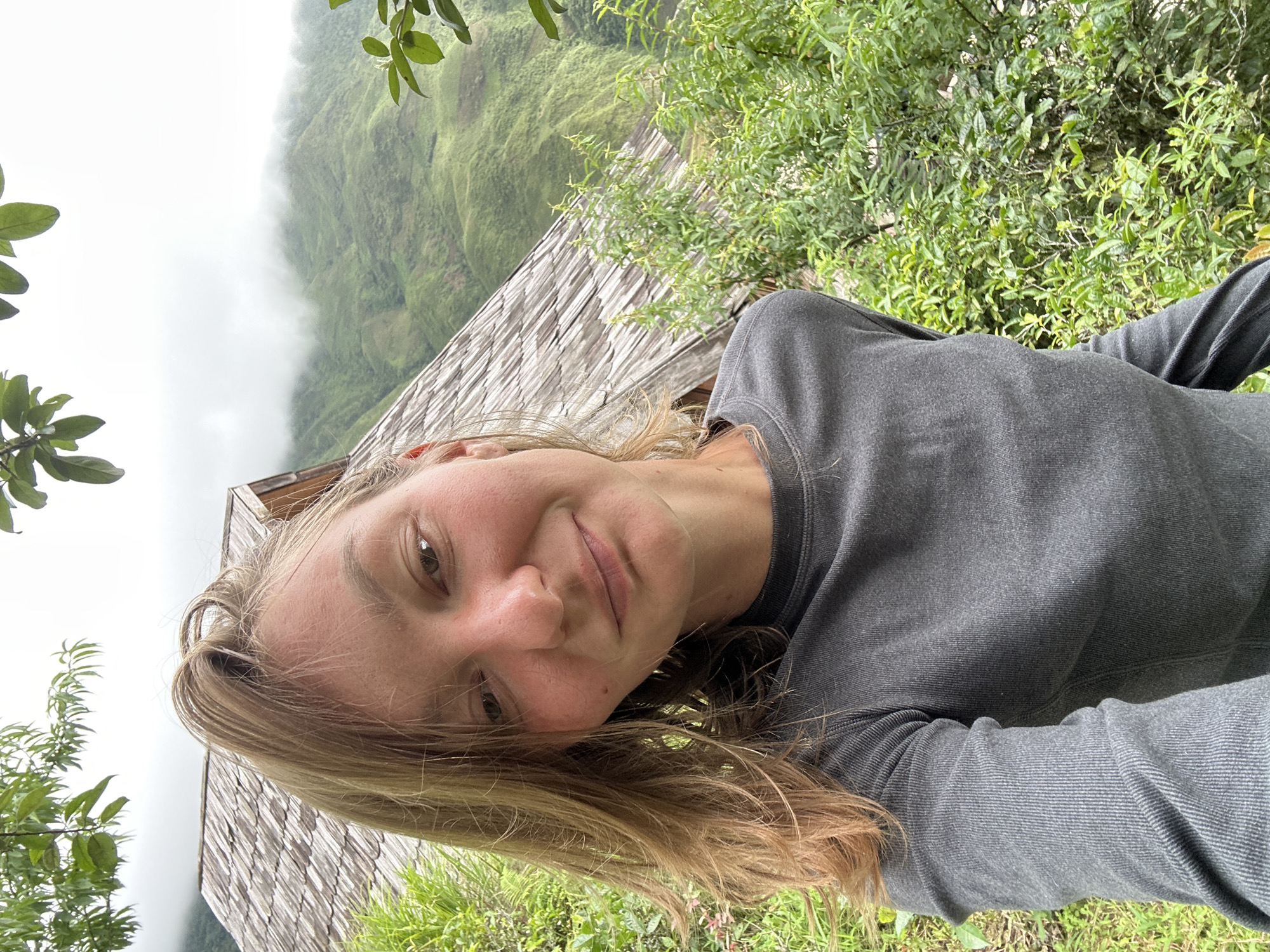 A young Caucasian woman with blonde hair and fair skin takes a selfie outdoors. She is wearing a dark grey long-sleeved shirt. Behind her, a wooden structure with a shingled roof is visible, adjacent to lush green foliage. In the background, rolling green hills are shrouded in mist under an overcast sky. The scene is set in Ta Xua, Vietnam, characterized by its mountainous terrain and natural beauty.