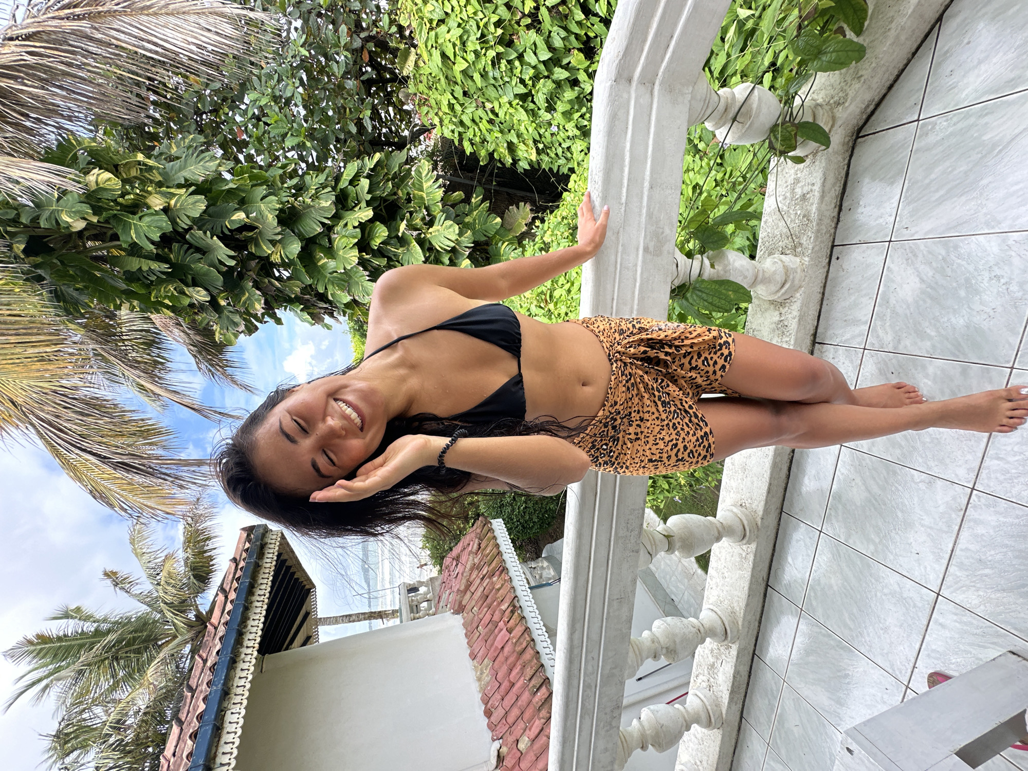 A young woman stands on a tiled balcony, leaning on a white ornate railing. She wears a black bikini top and a leopard-print sarong. Her eyes are closed, and she smiles, with her right hand touching her forehead. Palm trees and lush green foliage are visible behind her, with a glimpse of the ocean and sky in the background. The balcony appears to be part of a building with a red-tiled roof. The location is identified as Peleana, Sri Lanka.