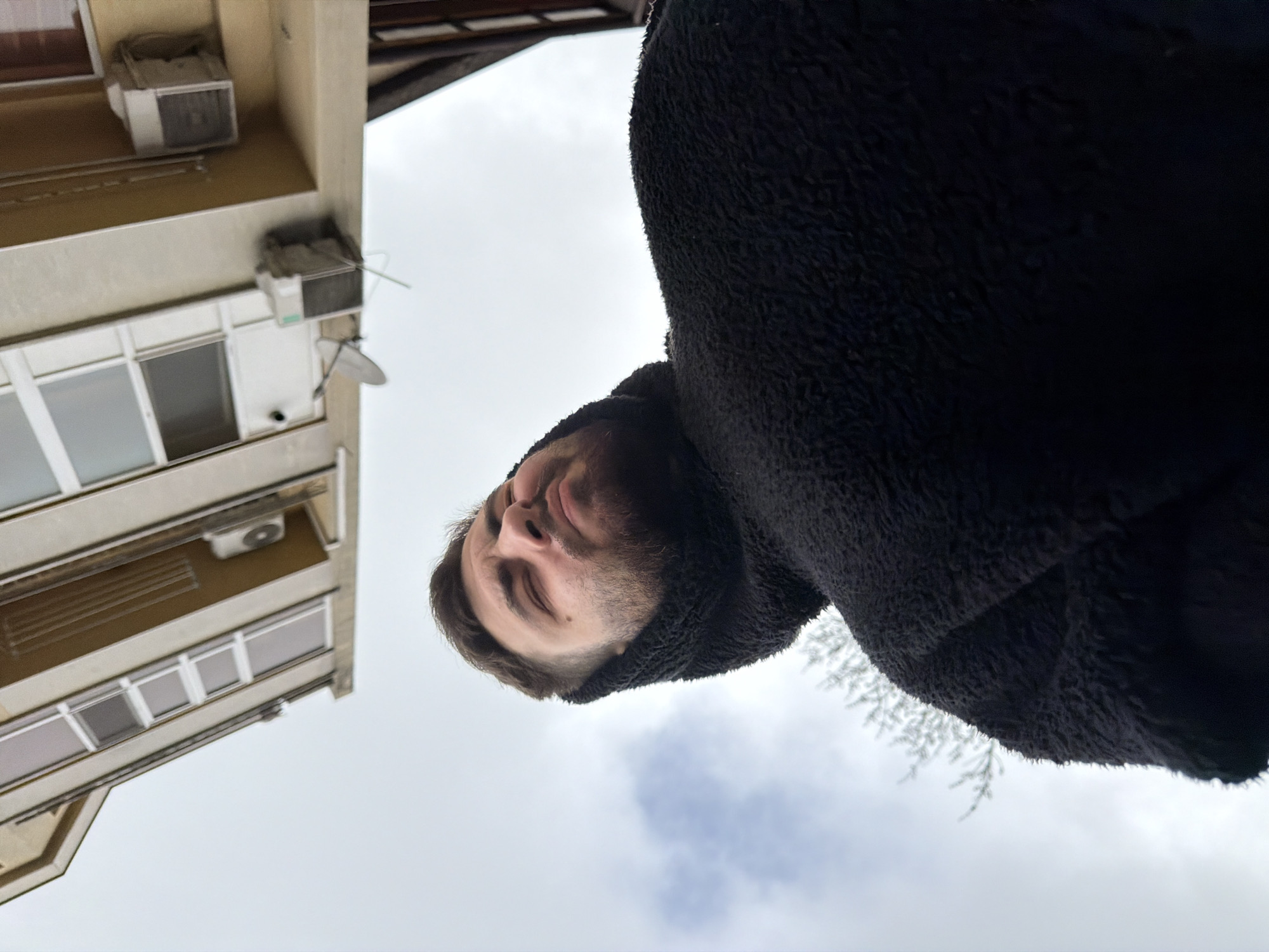 A low-angle photograph shows a man wearing a black, fuzzy hooded jacket. The sky, which is mostly cloudy with patches of blue, is in the background. A building with windows and air conditioning units is visible on the left side of the frame. Some branches appear to be frosted and are near the hooded part of the jacket. The location is Varna, Bulgaria.