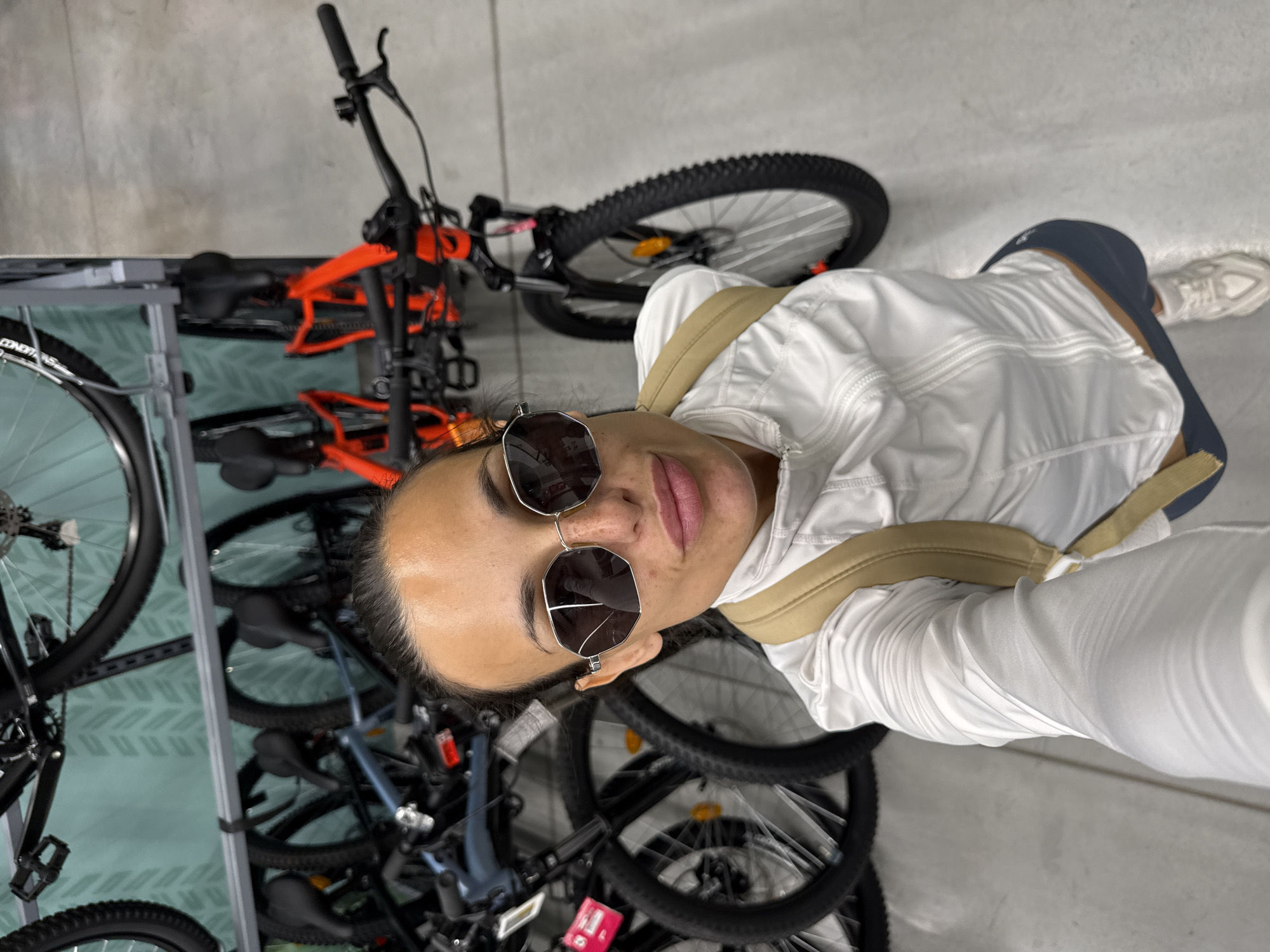 A woman is taking a selfie in a store that sells bicycles. She is wearing a white jacket, dark shorts, and white sneakers. She has on large, octagonal sunglasses. Behind her, several bicycles are mounted on a wall rack. The store has a concrete floor and a teal wall with a chevron pattern.
