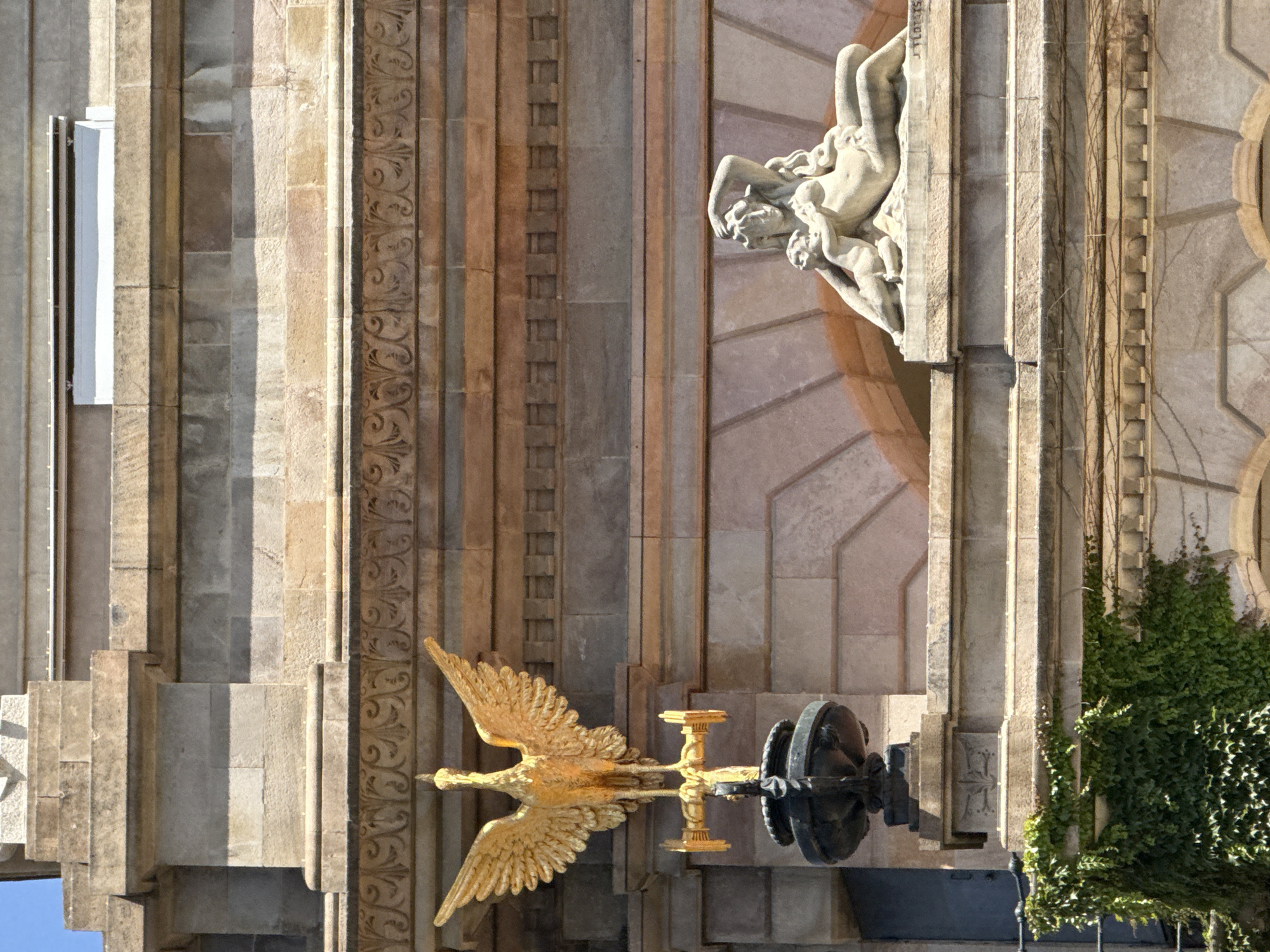 The image shows a close-up view of the exterior facade of a building in Barcelona, Spain. On the left, a golden sculpture of a bird with outstretched wings is mounted on a dark pedestal. The bird appears to be a phoenix, a mythical creature often associated with rebirth and fire. The sculpture is positioned against a wall constructed of large, rectangular stone blocks, with decorative horizontal bands of carved stone above and below.
To the right of the bird sculpture, on a slightly higher level, is a white stone sculpture depicting two figures. The dominant figure is a reclining woman with long, wavy hair. She is leaning back, with one arm raised and resting on her head, and the other arm extended. A smaller figure, possibly a child, is nestled close to her, with its arms around her. Both figures appear to be nude. The woman's form is rendered with soft, flowing lines.
The building itself is constructed of stone, with variations in color and texture indicating different types of stone and possible age or weathering. The architectural style suggests a classical or neoclassical influence, with decorative elements like moldings and carved friezes. Green ivy is visible growing on the lower left portion of the facade, adding a touch of nature to the stone structure. The sky is visible in the upper left corner, suggesting it is daytime. The overall impression is one of grandeur and artistry, typical of public buildings or monuments in historic cities.