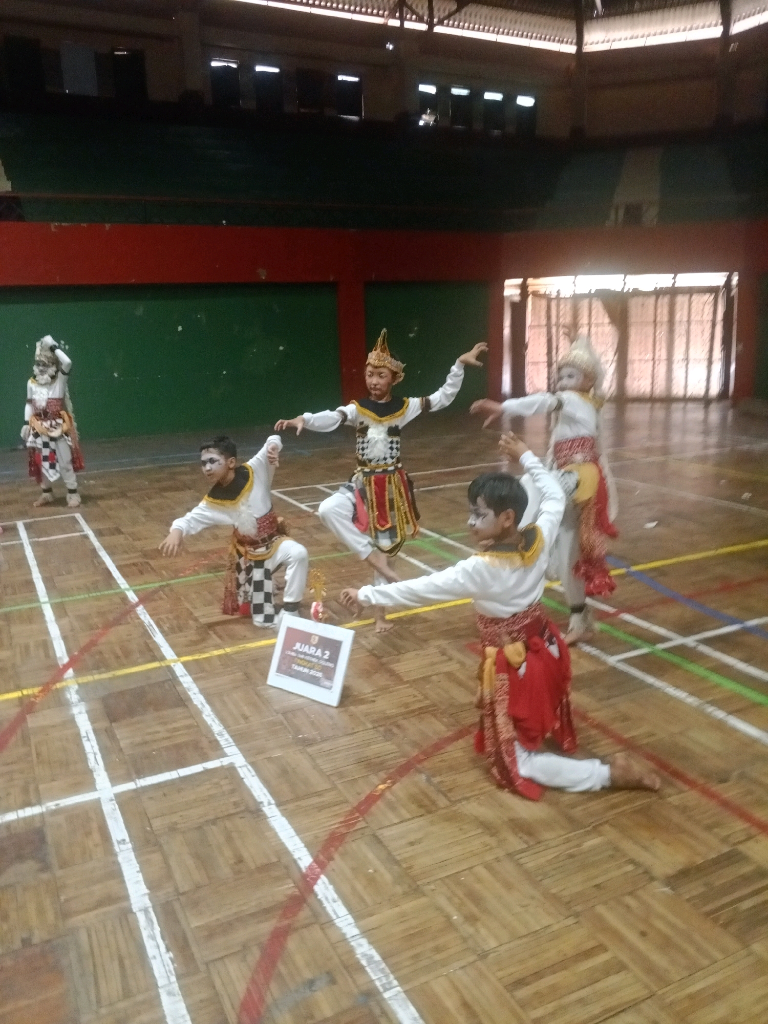 The image depicts a group of children dressed in traditional Balinese attire performing a dance on a wooden floor in what appears to be an indoor sports hall. The hall has green walls and elevated seating areas. The children are wearing ornate headdresses, colorful costumes with checkered patterns, and face paint. One child in the foreground is kneeling with arms outstretched, while another is in a crouching pose. In the background, two other children are standing, also in costume. A "JUARA 2" (2nd Place) award is visible on the floor in the middle ground. The lighting suggests an indoor environment, possibly during an event or competition. The scene is likely in Wuryorejo, Indonesia, given the context of traditional Indonesian dance performances.