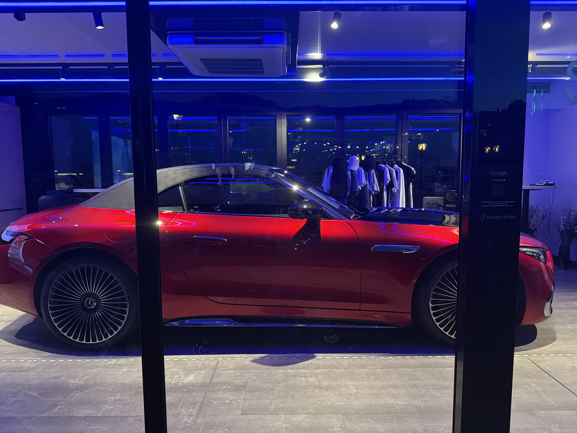 The image shows a red convertible car displayed indoors, likely in a showroom. The car is a Mercedes-Benz SL with the top down, revealing a black soft top. It is parked on a tiled floor, with a black pillar obscuring part of the view. The background features large windows with a city skyline visible through them, illuminated by streetlights and building lights. There are also clothes on racks in the background, suggesting the space might be a boutique or a multi-purpose venue. Blue LED lighting is prominent throughout the scene, both above the car and reflected on its surface. A sign with the "Google" logo and stars is visible on the black pillar, as is the "SILVER STAR" logo. The car is the main subject, and it is presented in a polished and well-lit manner. The overall impression is one of a modern and sophisticated commercial space. The location in Sveti Vlas, Bulgaria, is an important contextual detail, indicating a specific geographical setting for this display.