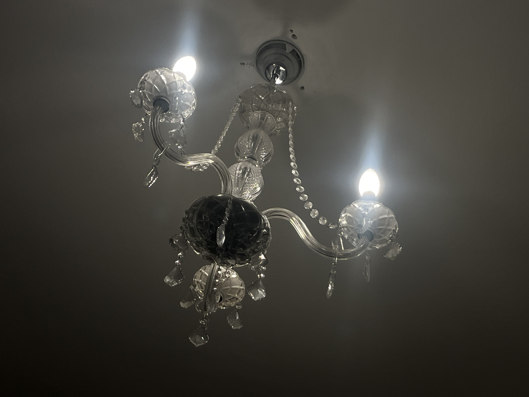 The image shows a chandelier hanging from a ceiling. The chandelier has multiple arms extending from a central point, each terminating in a lightbulb. The chandelier is adorned with crystal-like elements that dangle from the arms and central structure. The ceiling appears to be plain and relatively featureless. There are no visible people, actions, or interactions in the image. It is not possible to confirm that the location is in Barcelona, Spain, from the visual information provided.