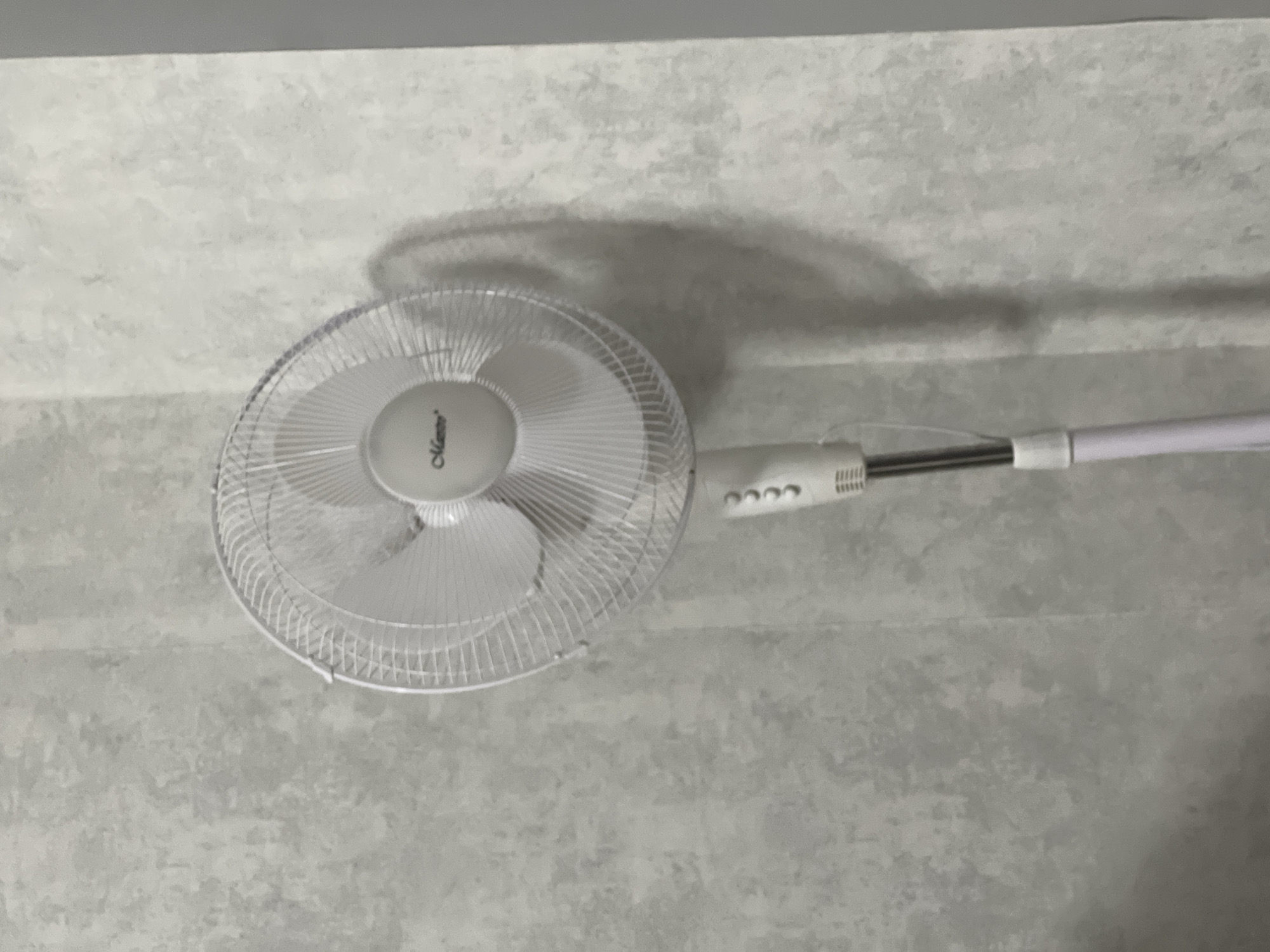 A white oscillating fan with a brand name "Maestro" is positioned against a textured grey wall. The fan has a circular grille with thin, vertical metal bars and a central circular housing. It is mounted on a white adjustable stand with a visible power cord. Below the fan head, there is a control panel with several buttons and a ribbed section on the stand. The background is a plain, textured grey wall with subtle variations in tone. The lighting suggests an indoor environment, casting a soft shadow of the fan on the wall to its right. There are no people present in the image. The scene appears to be a typical indoor setting.