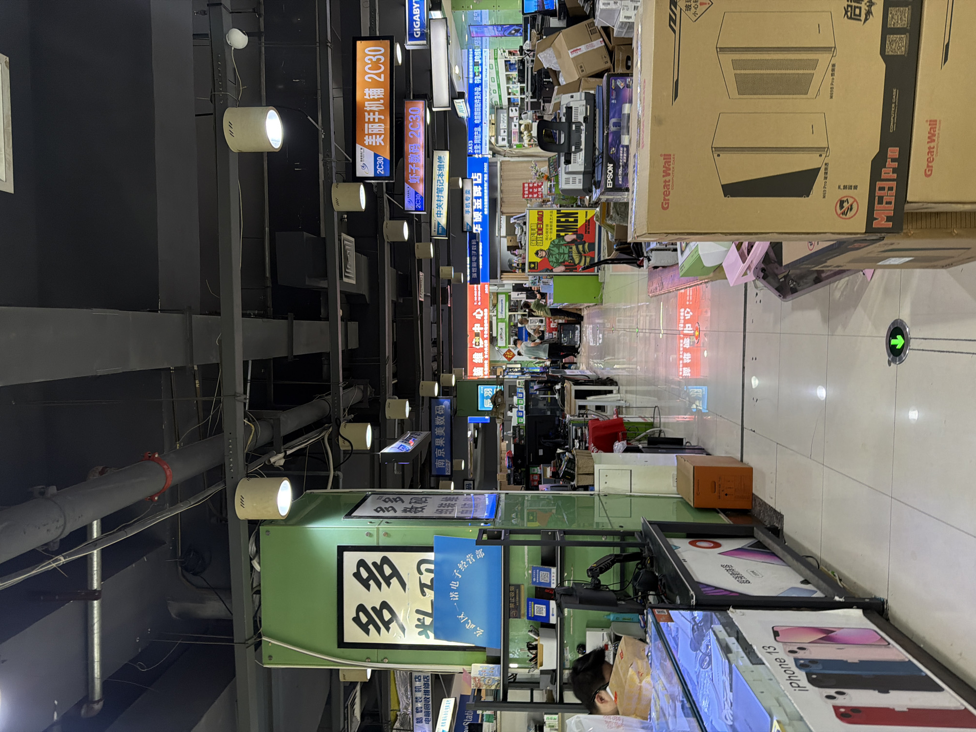 This is an indoor shot, likely inside a computer and electronics market or mall. The perspective is looking down a narrow aisle lined with various vendor stalls. Banners and signage hang overhead, with Chinese characters and English text visible. One banner advertises "Meili Mobile Phone Shop 2C30" (美丽手机铺 2C30) and another mentions "Zhongguancun Laptop Repair". Other store signs include "Duodu" (多多) and "NanJing Guomei Digital" (南京果美数码).
Several boxes, including one for a "Great Wall M69 Pro" computer case, are stacked in the aisle. The environment is cluttered with electronics, computer parts, and packing materials. There are some individuals visible in the background and a person wearing a mask and looking at a phone on the right side of the image.
The location is confirmed by one of the vendor signs, which includes the phrase "Xuanwu District Yinuo Electronics Operating Department" (玄武区一诺电子经营部), indicating it is within Xuanwu District in China. The lighting is a mix of fluorescent overhead lights and spots.