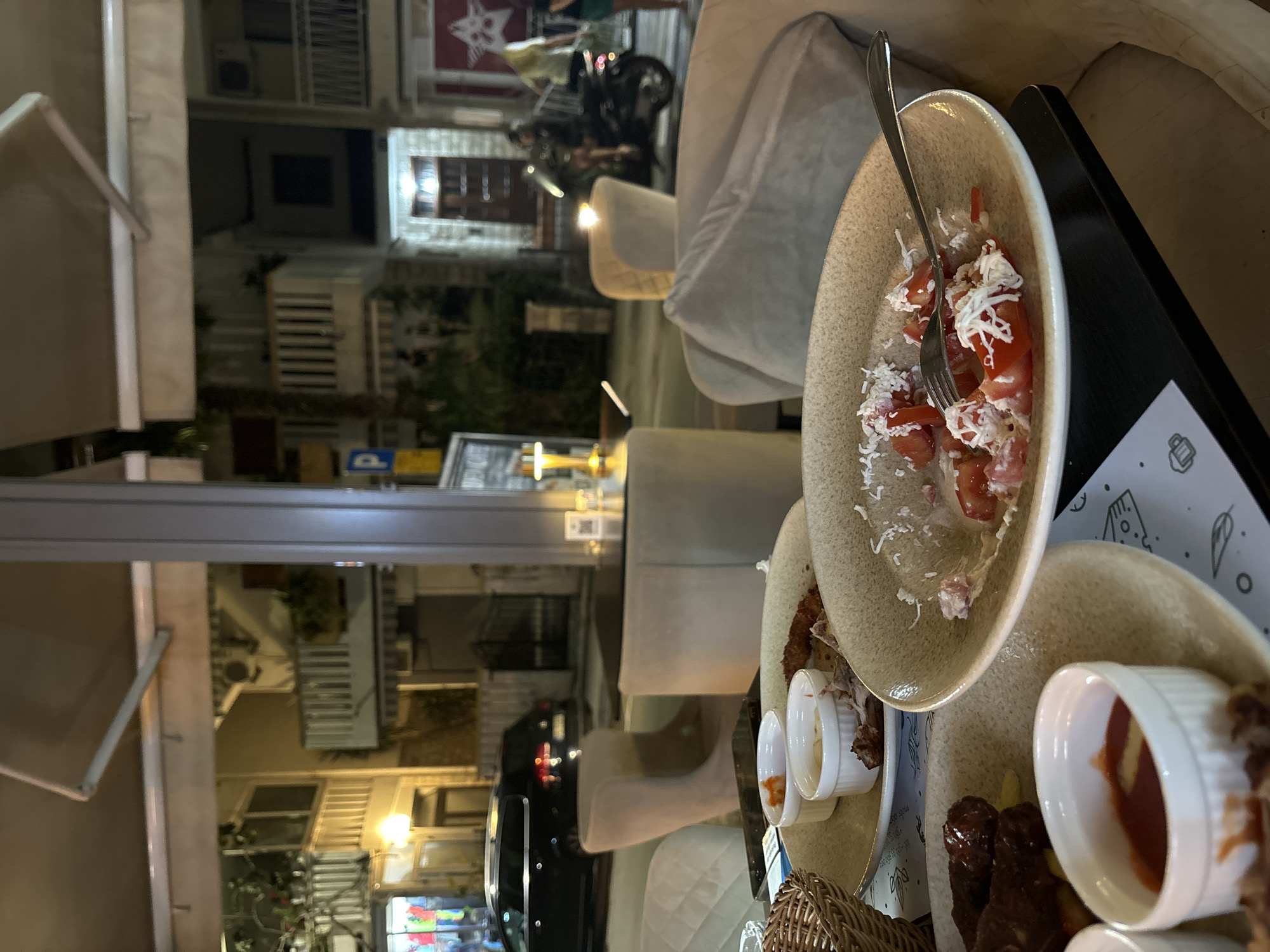 A dimly lit outdoor dining scene at night in Budva, Montenegro, shows the remnants of a meal on a table. In the foreground, a plate contains a salad of tomatoes and cheese with a fork. Another plate holds what appear to be sausages and sauce. Behind these plates, several white ramekins are visible, some containing dips or sauces, and a basket. The background reveals a street scene with buildings illuminated by artificial lights. A black car is parked on the street, and a person on a scooter is seen in motion. The overall ambiance suggests a restaurant or cafe setting on a street in Budva.