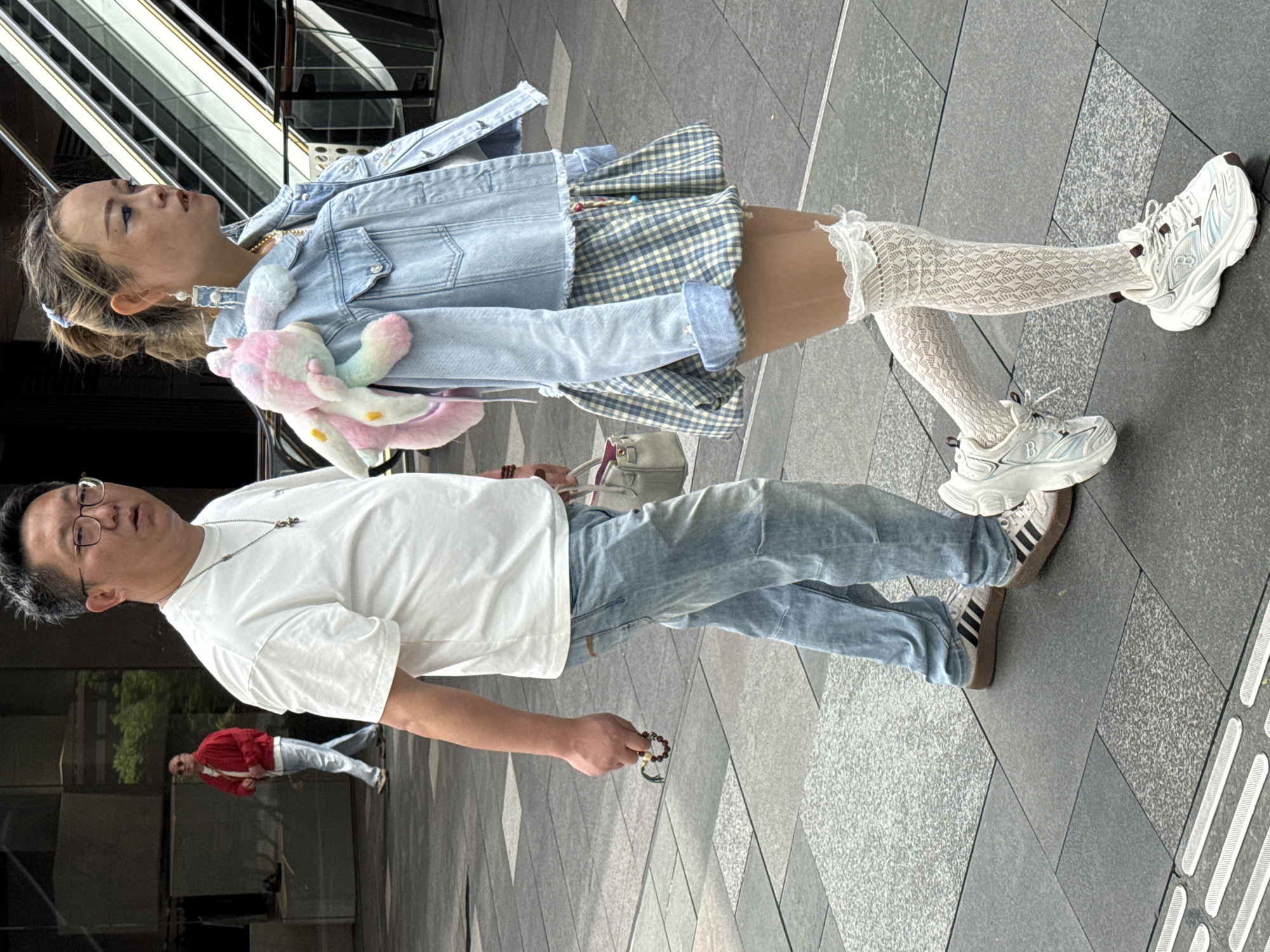 A man and a young woman are walking outdoors in Jinjiang District, China, on a gray paved walkway with a tactile paving indicator. The woman is positioned slightly ahead of the man, and both appear to be walking in the same direction.
The woman is wearing white sneakers, lace-patterned white knee-high socks, a blue and white checkered skirt, and a light blue denim jacket. She has a pastel-colored plush toy draped over her shoulder. Her hair is blonde and styled in pigtails.
The man is wearing a white t-shirt, light blue jeans, and dark sneakers with black stripes. He wears glasses and a necklace. He is holding a bracelet.
In the background, another man is visible in a red shirt and jeans. A glass building with an escalator is also visible in the background.