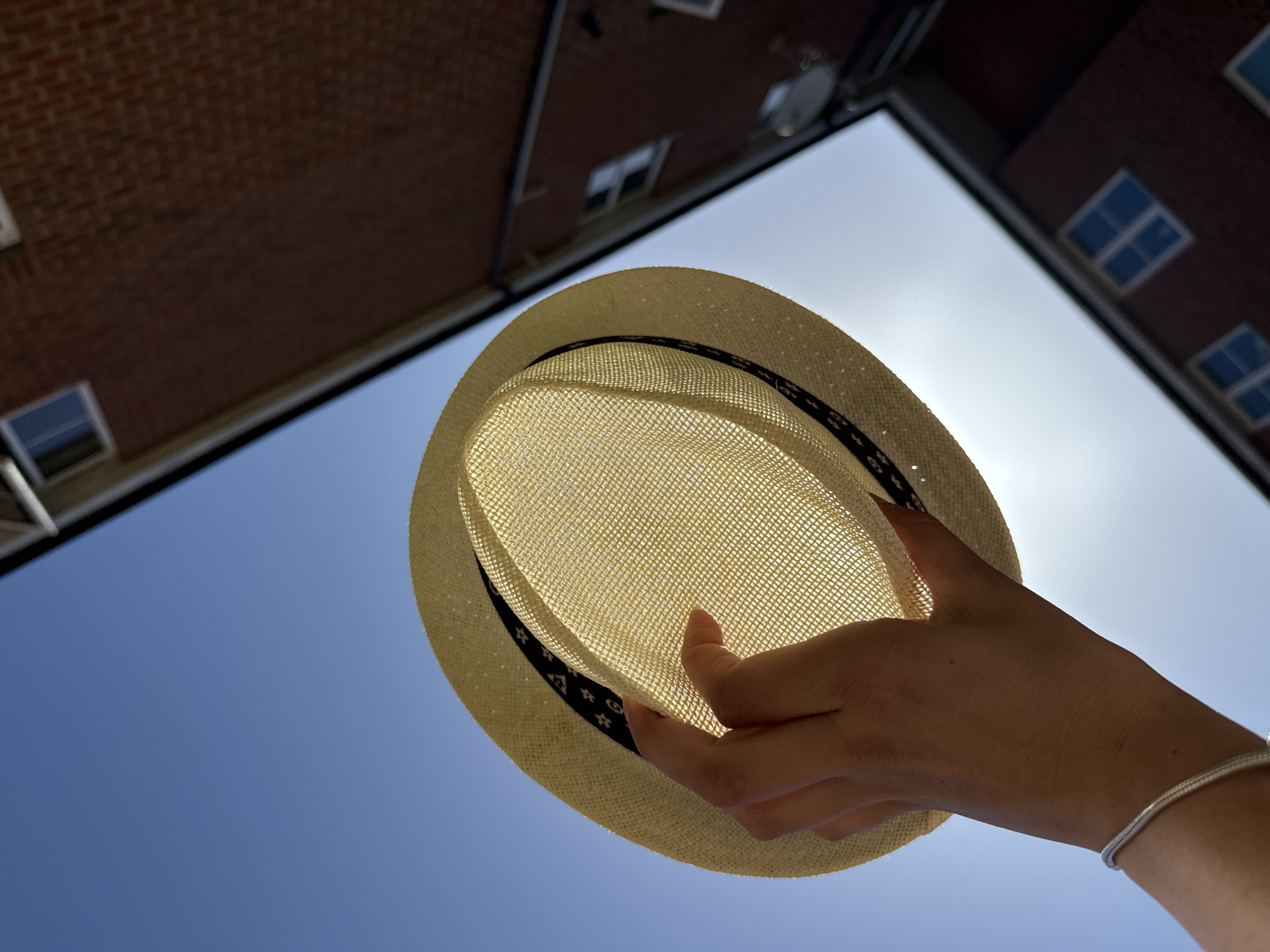 A low-angle shot shows a straw fedora hat held by a hand against a bright blue sky, partially obscuring the sun. The hat has a black patterned band around the brim. The hand is light-skinned and adorned with a silver bracelet. In the background, blurred brick buildings with windows flank the sky. The image appears to be taken in an urban environment, potentially Great Yarmouth, United Kingdom, due to the architectural style of the buildings.