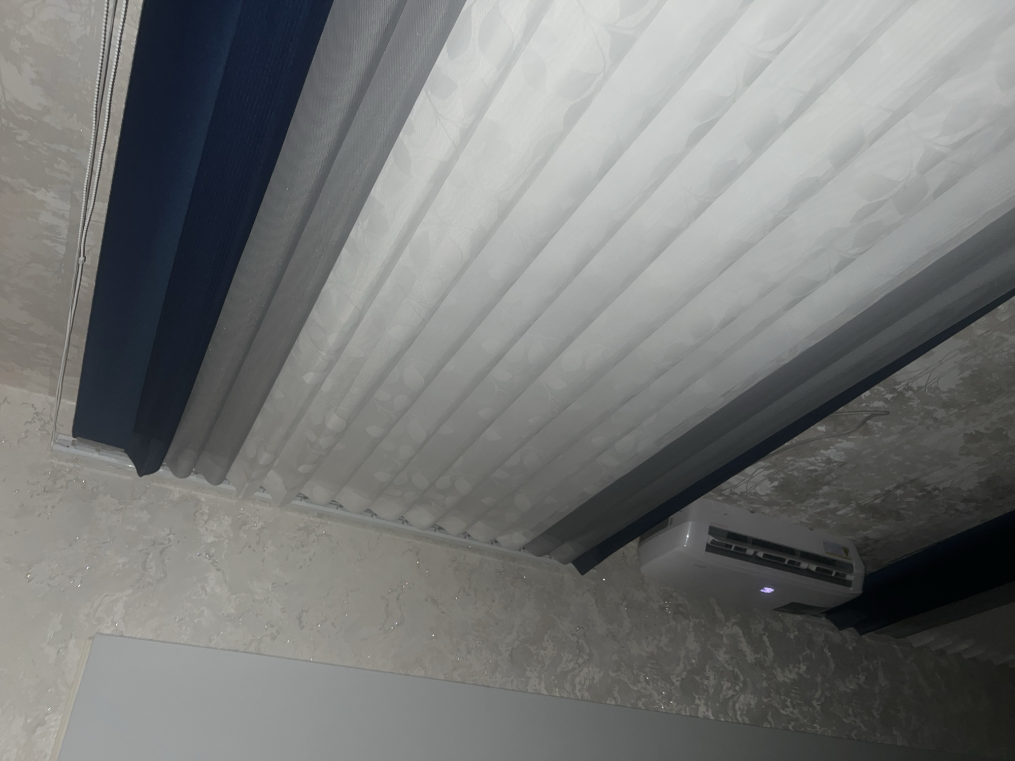 The image displays a close-up view of window blinds and an air conditioning unit mounted on a textured wall. The blinds are layered, featuring dark blue, grey, and white translucent fabric with a subtle floral pattern. The wall has a marble-like texture with flecks of glitter. A white air conditioning unit is visible on the wall below the blinds. The scene appears to be indoors, likely in a residential setting. There is no information available to determine the specific location within Yukari Chek, Uzbekistan.