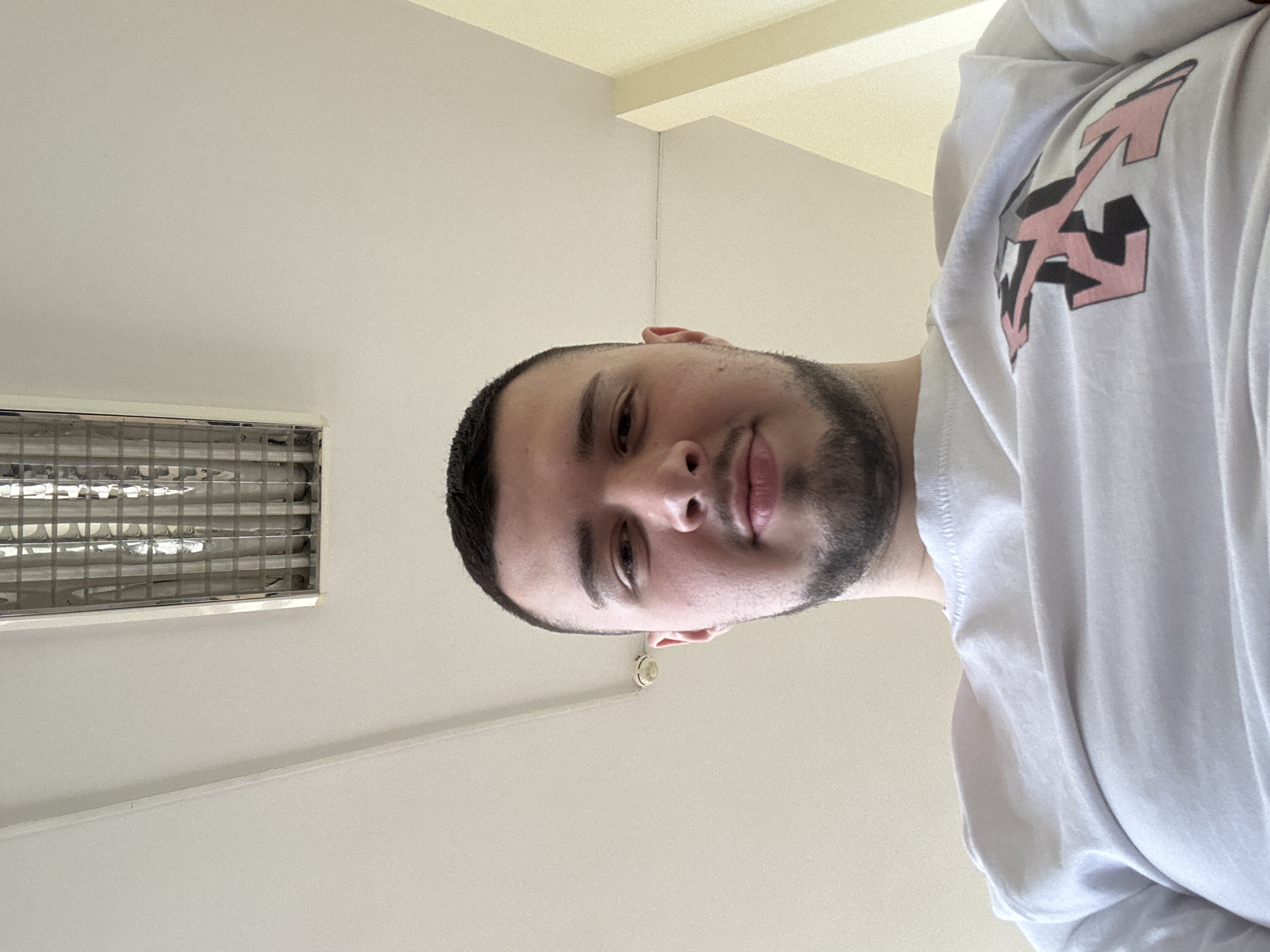 A man with a dark beard and a white t-shirt with a pink and black graphic stands in an indoor space. The ceiling is white with a rectangular fluorescent light fixture to the left. The specific location in Sofia, Bulgaria, cannot be determined from this image.