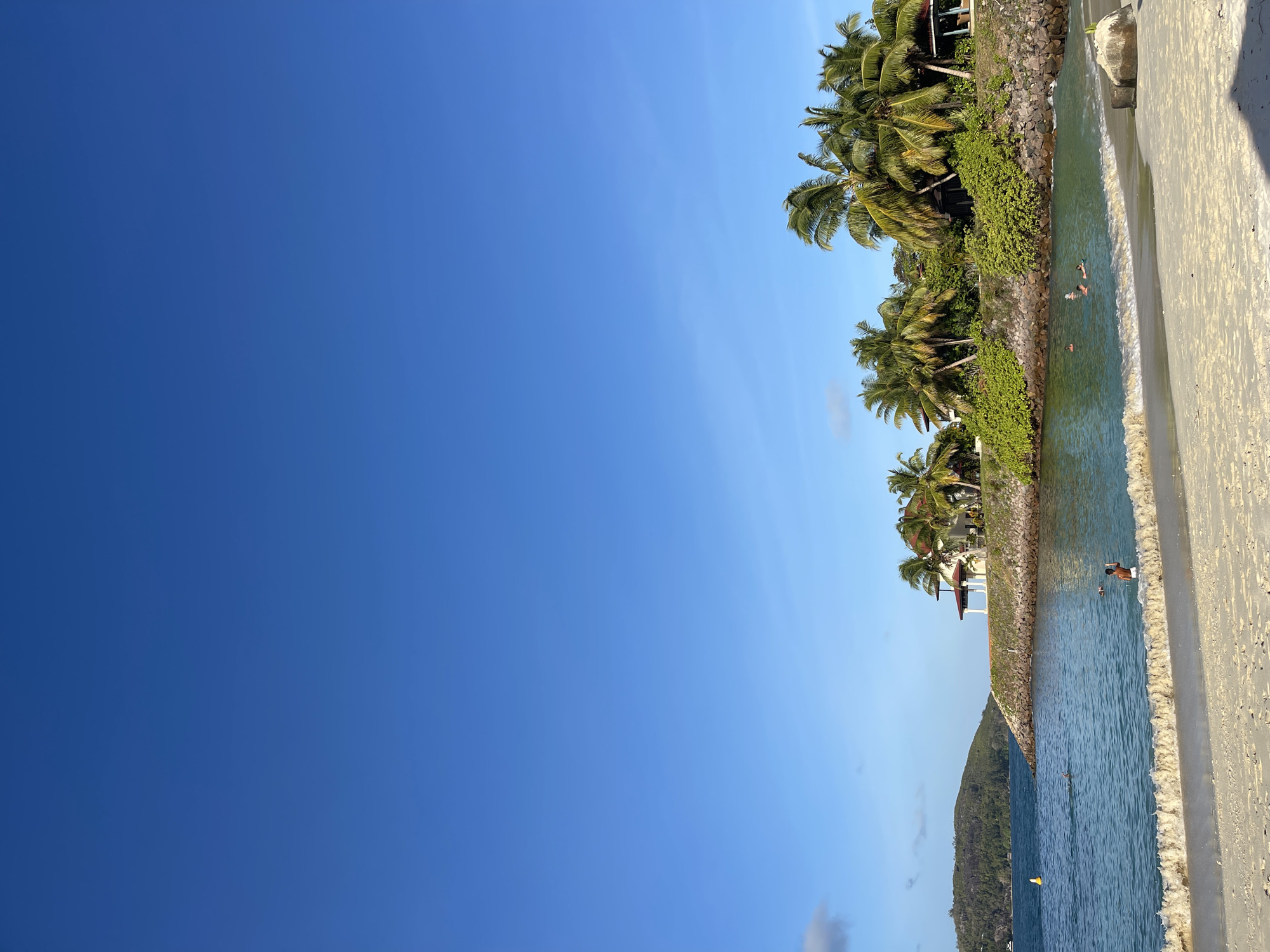 A tranquil tropical beach scene unfolds under a clear, vibrant blue sky. The foreground features a sandy shoreline with gentle waves lapping at the beach. In the shallow turquoise water, several people are visible swimming and enjoying the ocean. Further out, a yellow buoy floats, and a verdant, tree-covered island rises in the distance. The midground showcases a lush green landscape dotted with palm trees, overlooking a rocky shoreline. A building with a reddish roof and white columns is nestled amongst the greenery, suggesting a resort or residential area. The overall impression is one of a peaceful, idyllic coastal setting.