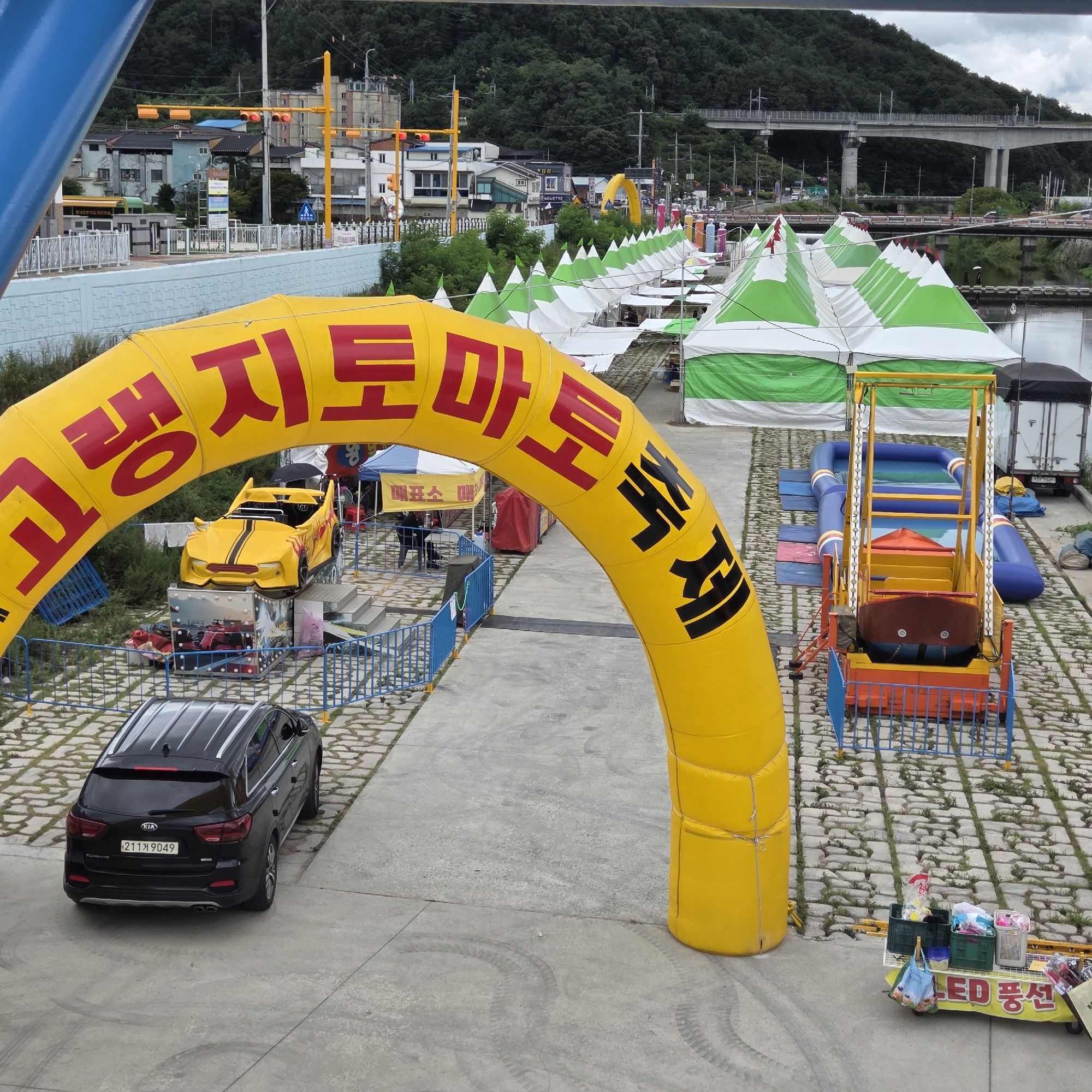 The image captures a street scene in Dunnae-myeon, South Korea, on a cloudy day. A large, inflatable yellow archway with Korean text written in red and black characters is a prominent feature, spanning across a pathway. Beneath the archway, a small, yellow electric car ride designed for children is parked, with a metal structure behind it. To the right of the car ride, a small inflatable water slide with blue and green tubing is visible. In the background, several white and green tents are set up in rows, suggesting an outdoor market or event. A black Kia Sorento SUV is parked on the left side of the pathway, facing away from the camera. The ground is paved with interlocking stones, with grass growing in the gaps. Further in the background, buildings, trees, and an elevated highway bridge are discernible. A cart selling LED balloons is visible on the right side of the frame, with various items displayed in baskets.