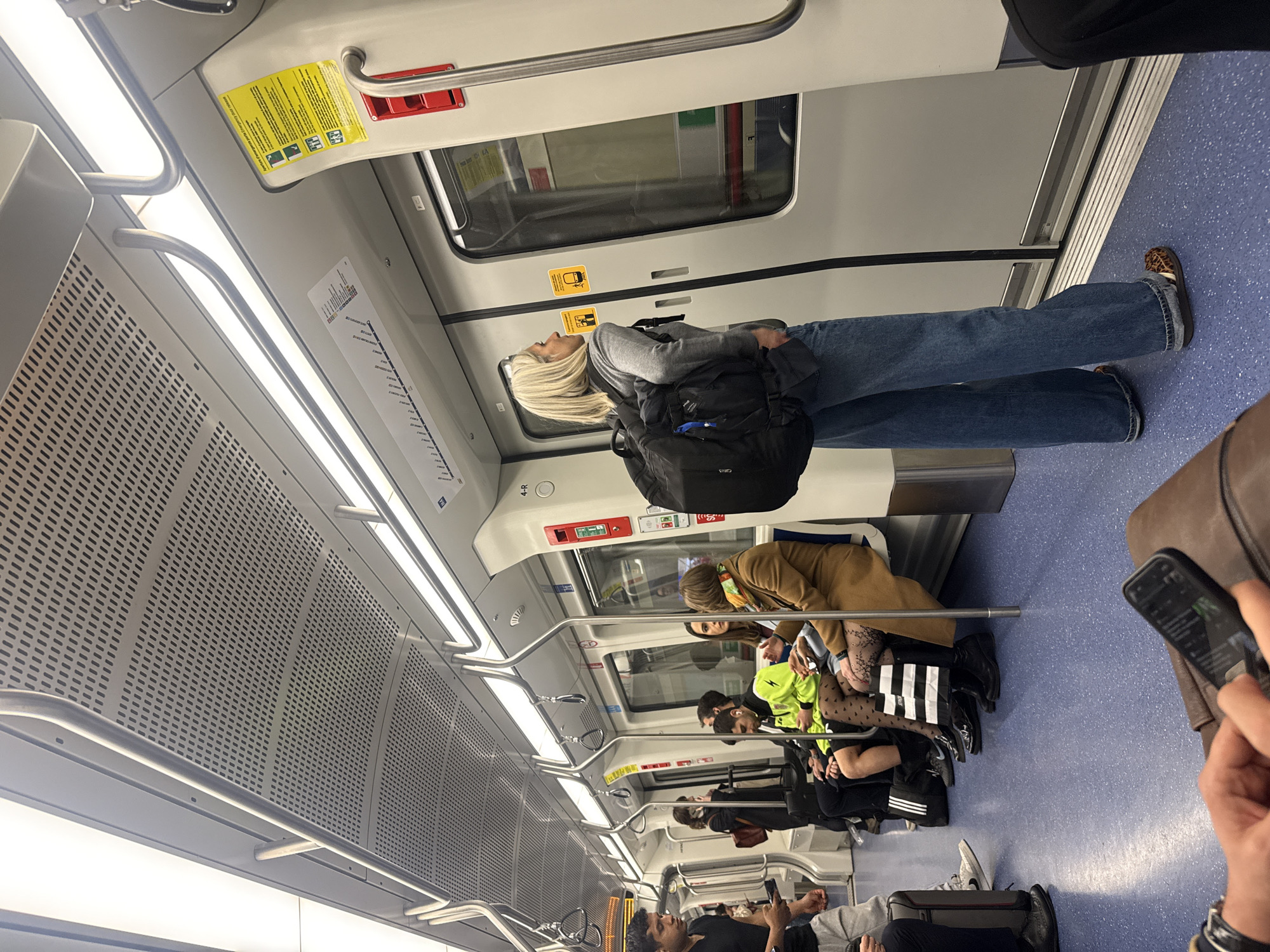An overhead view captures the interior of a modern train or subway car, rotated 90 degrees clockwise from its natural orientation. The car features a clean, light grey and white color scheme with a blue, textured floor. Bright LED strip lighting runs along the ceiling, which also has perforated acoustic panels. Silver handrails are visible, both horizontal bars above the seats and vertical poles for standing passengers.
In the foreground, a blonde woman with long hair stands, looking towards the front of the train. She wears a grey hoodie, loose-fitting blue jeans, and leopard-print flat shoes. A black backpack is on her back.
Further down the car, several other passengers are seated on what appear to be bench-style seats. One woman with brown hair wears a brown coat and patterned black tights, and a black and white striped shopping bag is visible near her feet. Another person seated nearby wears a bright green top. Various other passengers are visible further back, many engaged with their mobile phones. One man in a black t-shirt holds a phone.
Visible objects include emergency pull handles or buttons (red) mounted above the windows, and windows themselves reflecting the interior. A yellow sign near the ceiling contains pictograms for accessibility and safety, along with dense text, partially readable as "Achtung!" (German for "Attention!"). Another small diagrammatic map of a train line is visible on a wall panel. The overall setting is brightly lit, indicating a modern public transport system.