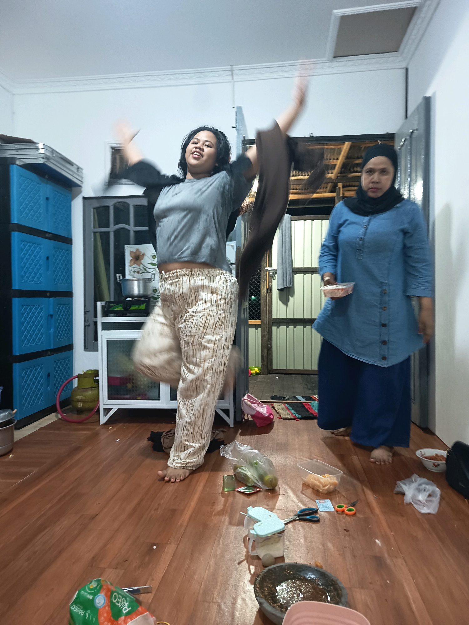 In an indoor residential setting, likely a kitchen or common area, two individuals are present. The primary subject, a young woman, is captured mid-jump or dance, with her arms outstretched and a dynamic pose. She wears a grey t-shirt and patterned trousers. To her right, a woman in a blue denim-like shirt and dark headscarf stands holding a small container of food. The room features a wooden floor, blue plastic storage cabinets on the left, a small white trolley with a gas cylinder and cooking utensils, and a doorway in the background leading to an outdoor or less finished area. Various items are scattered on the floor, including plastic bags, a bowl, scissors, and a mortar and pestle, suggesting recent activity or preparation. The lighting appears to be from overhead fixtures. The overall impression is of a candid, informal moment within a home. Based on visual cues, this scene is consistent with a residential interior in Banjarmasin, Indonesia.