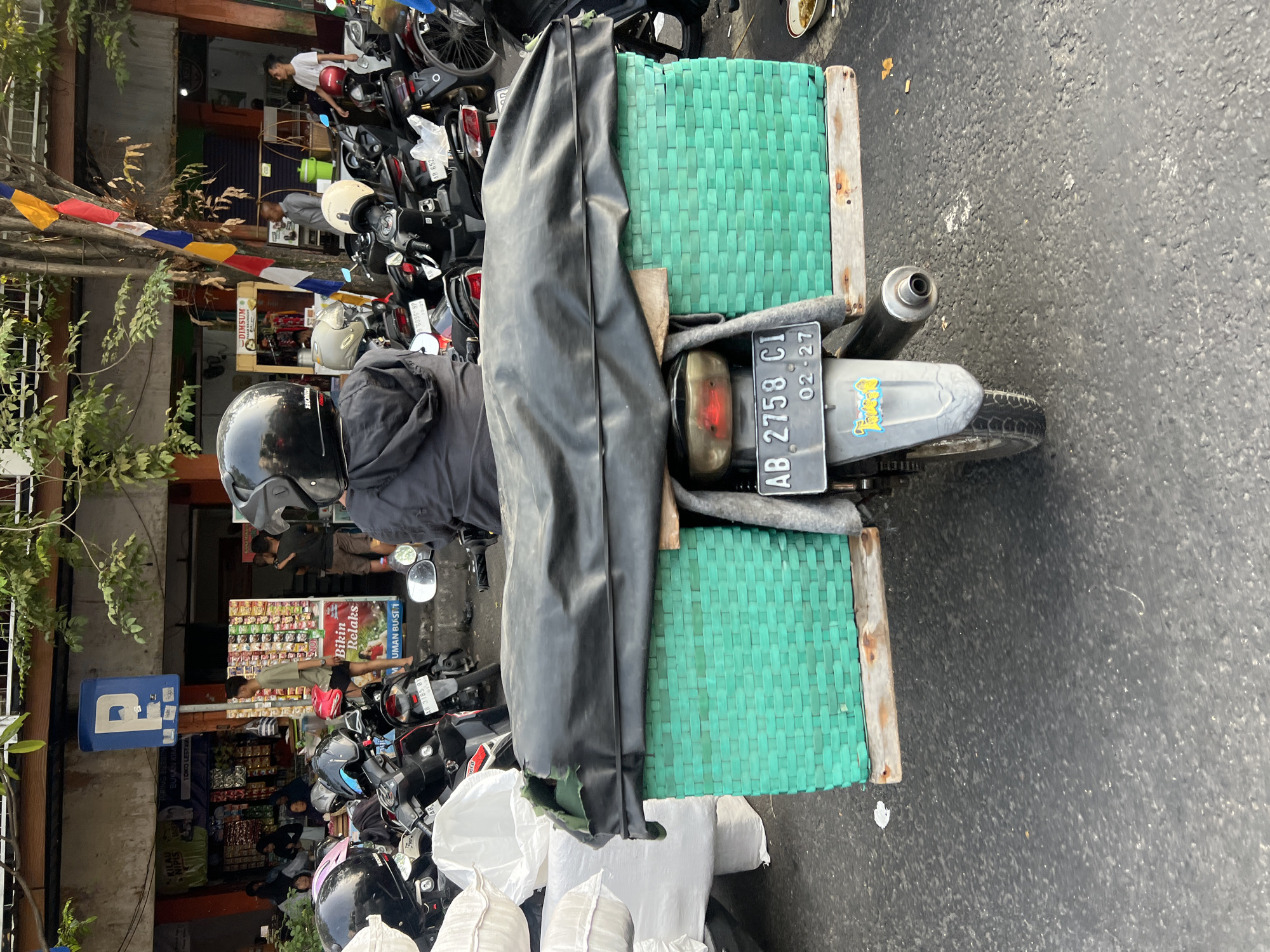 A rear view of a motorcycle with a large green woven basket attached to its back, covered by a black tarp, is parked on a street in the city of Yogyakarta, Indonesia. The motorcycle is positioned in the foreground, with its license plate reading "AB 2758 CI". A person wearing a black helmet is seated on the motorcycle, facing away from the camera. Behind the motorcycle, other parked motorcycles are visible, along with a storefront featuring various merchandise and signage. The environment appears to be an outdoor market or commercial area, with trees and flags overhead. The ground is paved with asphalt, and there are some discarded items and debris scattered around.