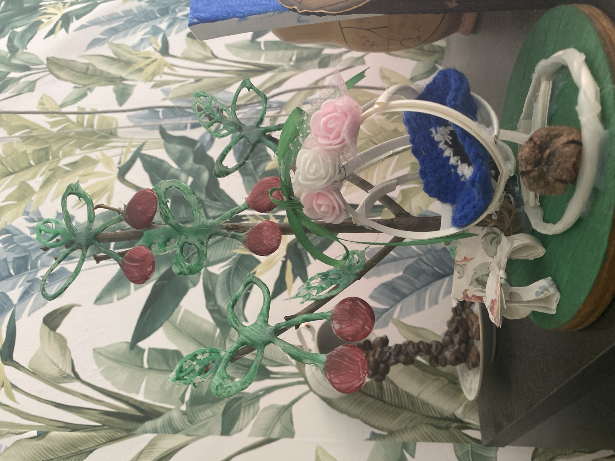 A miniature diorama is displayed against a backdrop of tropical-themed wallpaper. The diorama features a stylized green tree with red fruit, resembling cherries. Attached to the tree are small, green figures made from what appears to be a flexible material, with two of them holding pairs of the red fruit. There is a small white swing with a blue knitted cushion and a delicate white headpiece adorned with pink and white fabric roses. To the left of the swing, a miniature bench and table set, decorated with a floral pattern, is visible. In the background, a pile of dark brown coffee beans rests on a white plate. The diorama is set on a dark surface, likely a shelf or table, which is placed against the wall with the wallpaper. To the right of the diorama, a portion of a wooden object with painted details and a blue element is visible, possibly part of another decorative item or artwork. The lighting suggests an indoor setting, possibly a room in a home. The image is taken from a slightly elevated angle, looking down at the diorama. The scene does not appear to be located in Blagoevgrad, Bulgaria, as there are no specific indicators of that location.