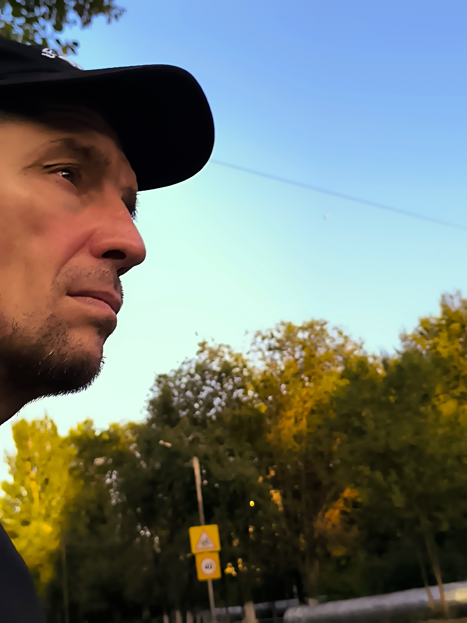 An eye-level, medium shot captures a man in profile, wearing a black baseball cap and a dark shirt. He is looking to the left of the frame, with his face partially obscured by the cap's brim. The background consists of a clear blue sky and out-of-focus trees with yellow and green foliage, suggesting an outdoor setting possibly in Qarağandı city administration, Kazakhstan, during autumn. A thin wire or line is visible diagonally across the sky.