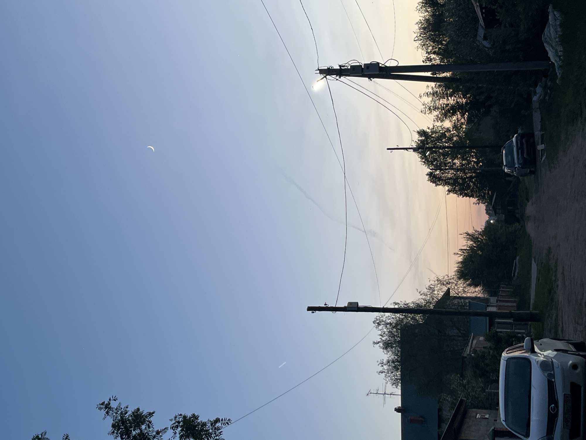 The image shows a street in what appears to be a residential area, possibly in Chaikovsky, Russia, although there is no definitive evidence to confirm this location. The image is taken at dusk or dawn. The sky transitions from blue to a pale orange/pink hue near the horizon. A crescent moon is visible in the upper left portion of the sky. Several utility poles are present, with electrical wires crisscrossing the sky. The poles run along a narrow dirt road. Vegetation, including trees and shrubs, lines both sides of the road, obscuring some houses. Two vehicles are visible. A white car is parked on the right side of the image, near the bottom of the frame. Another car, darker in color, is further down the road. There are no people visible in the scene. The overall impression is one of a quiet, rural street at twilight.