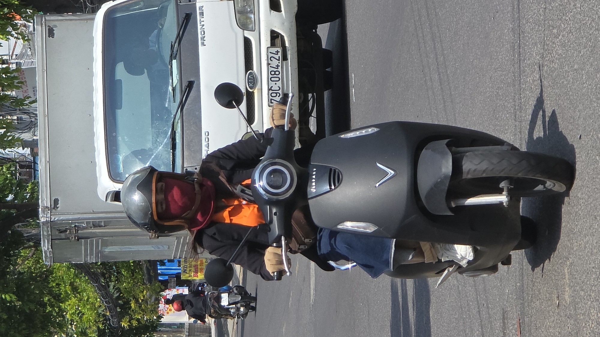 The image captures a street scene in Nha Trang, Vietnam. The primary subject is a person riding a black VINFAST electric scooter. The rider wears a grey helmet, an orange shirt, and a red face covering. A white KIA FRONTIER truck is positioned beside the scooter. The license plate number on the truck reads 79C-084.24. Other scooters and street greenery are visible in the background. The setting appears to be a typical urban street with visible road markings.