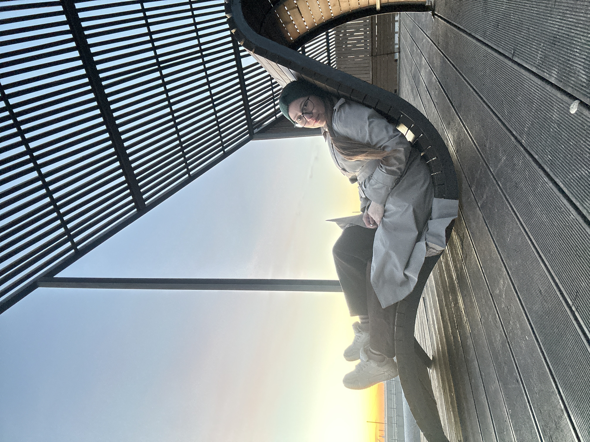 A woman wearing a gray coat, dark pants, white sneakers, a dark beanie, and glasses is sitting on a circular bench under a modern architectural structure. The structure consists of a series of parallel wooden slats overhead and a platform made of similar planks. The setting sun casts a warm light on the horizon, seen between the planks, suggesting the scene takes place outdoors.
While it is impossible to determine the location with certainty from the image alone, the architectural style and the light suggest that this could be somewhere in Saint Petersburg, Russia, given the request. The woman is holding something in her hands and appears to be reading.