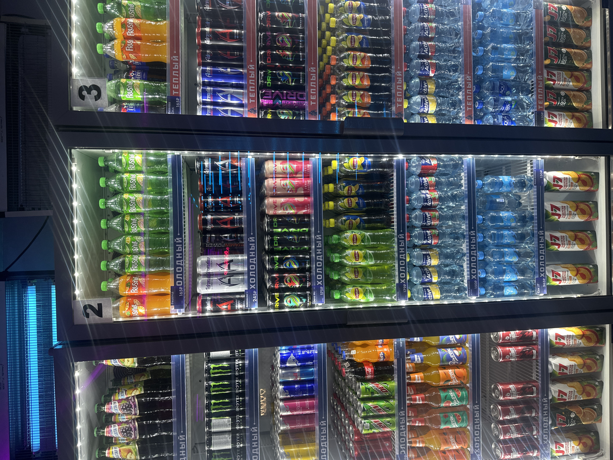 The image shows a commercial refrigerator filled with various beverages for sale. The setting is likely a retail store in Kazan, Russia, given the Cyrillic text on the product labels and price tags. The refrigerator has glass doors and interior lighting. The shelves are densely packed with bottled and canned drinks.
Displayed beverages include:
* "Frustyle" brand drinks in various flavors.
* "Adrenaline Rush" and "Drive Me" energy drinks.
* "Lipton" iced tea.
* "Aqua" mineral water.
* "J7" brand fruit juices.
* "Granat" juice.
* "Monster" energy drinks.
* "Red Bull" energy drinks.
* "Fanta" and "Mountain Dew" sodas.
* "Akhtari" brand drinks.
The refrigerator doors have numbers indicating their respective sections, labeled "2" and "3". Price tags are visible next to some products, and signage indicating "cold" (холодный) and "warm" (теплый) is present. The presence of a bug zapper with a blue light source is visible on the left side of the image, suggesting a location where insects are an issue.