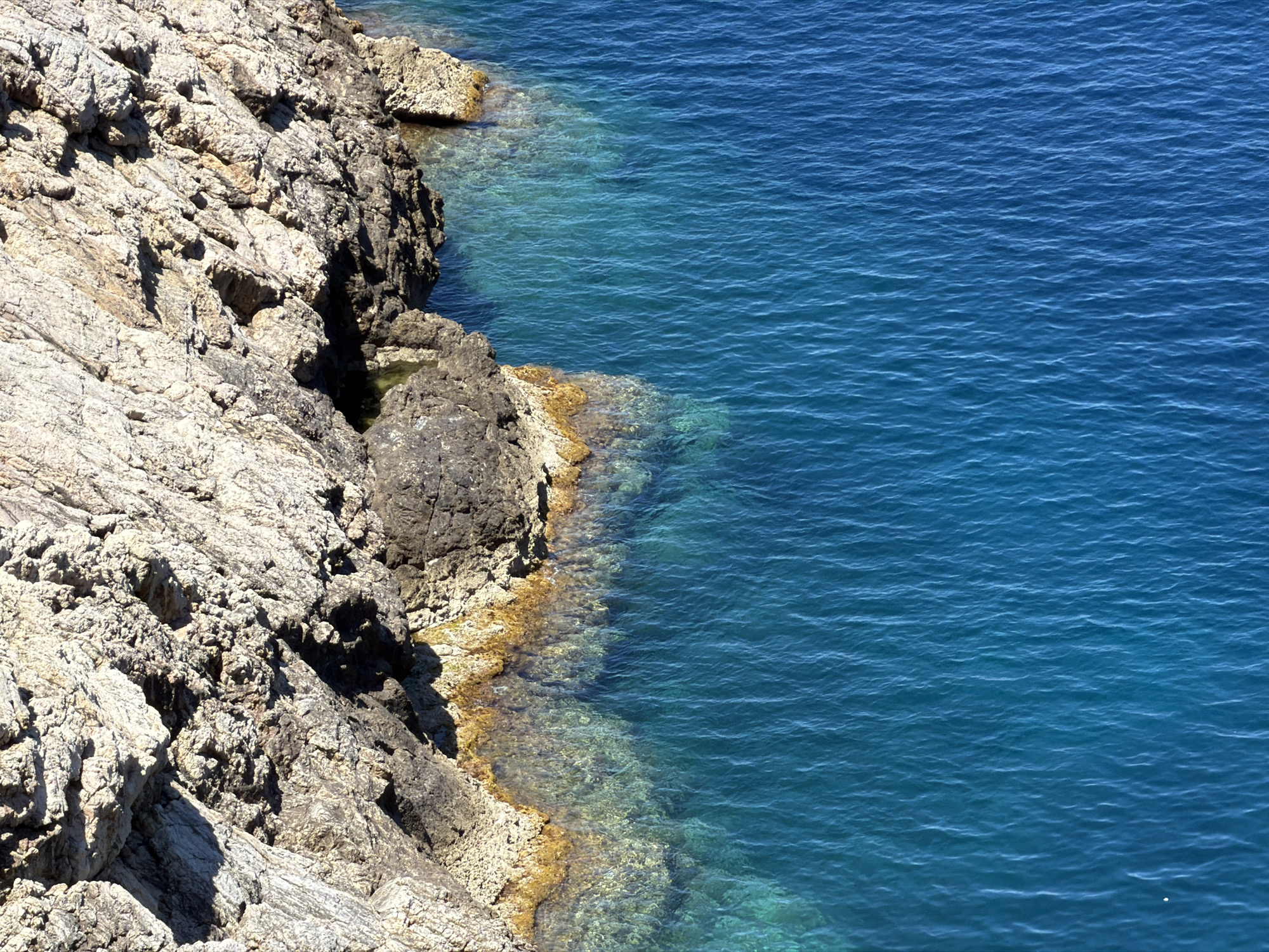 A high-angle, sunlit view shows a rocky coastline meeting the turquoise waters of the Mediterranean Sea in Capdepera, Spain. The rock formations are jagged and textured, appearing grey and tan, while the water near the shore is clearer, revealing the seabed. The ocean's surface transitions to a deeper blue further from the coast.