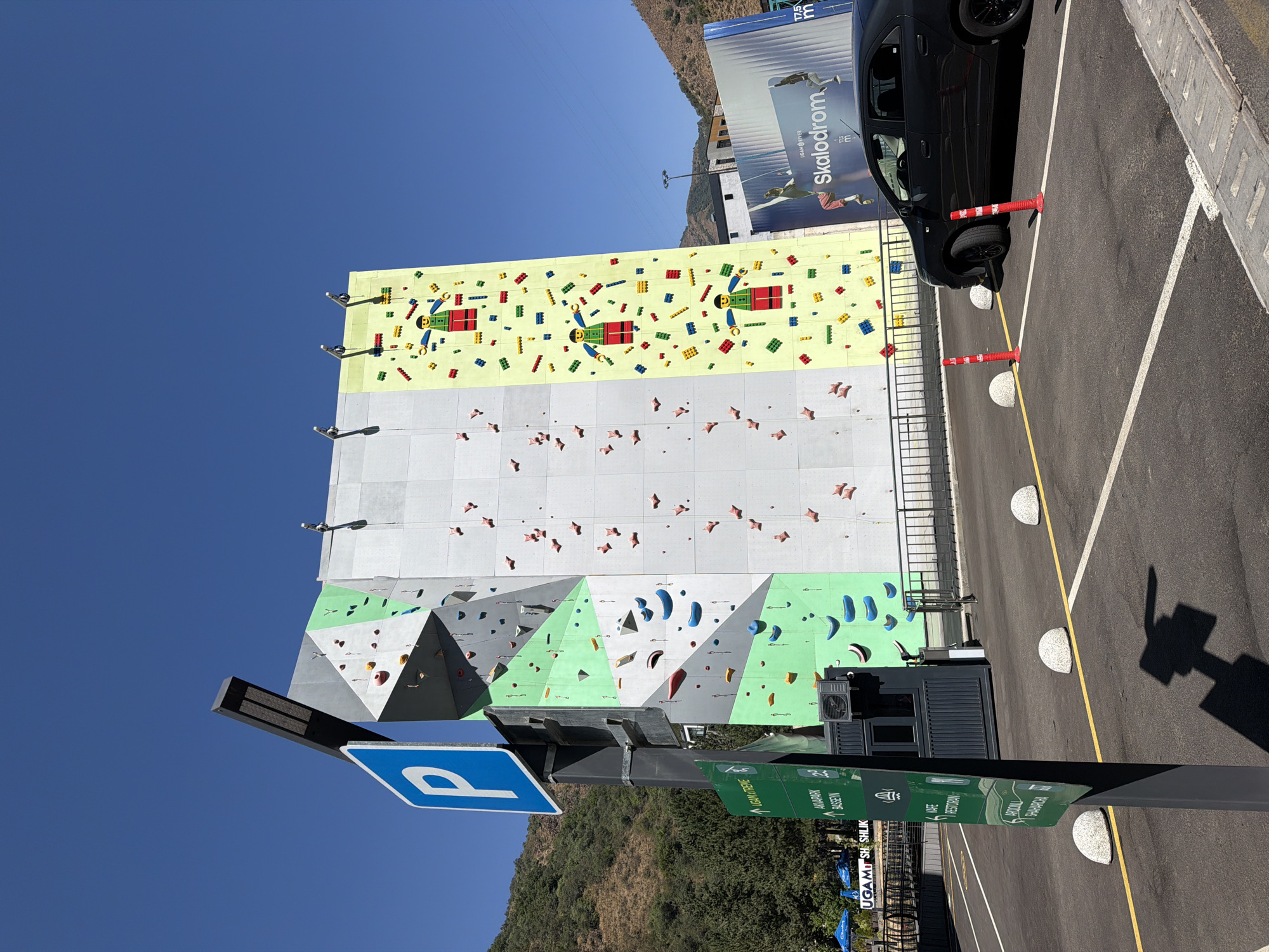 The image depicts a large, multi-colored climbing wall, known as a "Skalodrom," situated in Khumsan, Uzbekistan. The climbing wall is divided into several sections, with varying colors and climbing holds, and features a large graphic of cartoon figures resembling Lego characters on the rightmost section. To the left of the climbing wall, a blue and white parking sign is prominently displayed on a pole, accompanied by green directional signs. In the foreground, a black sedan is parked in a designated parking space, marked by white lines and bollards. Red traffic cones are also visible in the parking area. Behind the climbing wall, a billboard with the text "Skalodrom" and images of climbers is present. The background shows a clear blue sky, a green, tree-covered hillside, and some buildings. The overall scene appears to be a recreational area on a sunny day, with a focus on the climbing facility.
