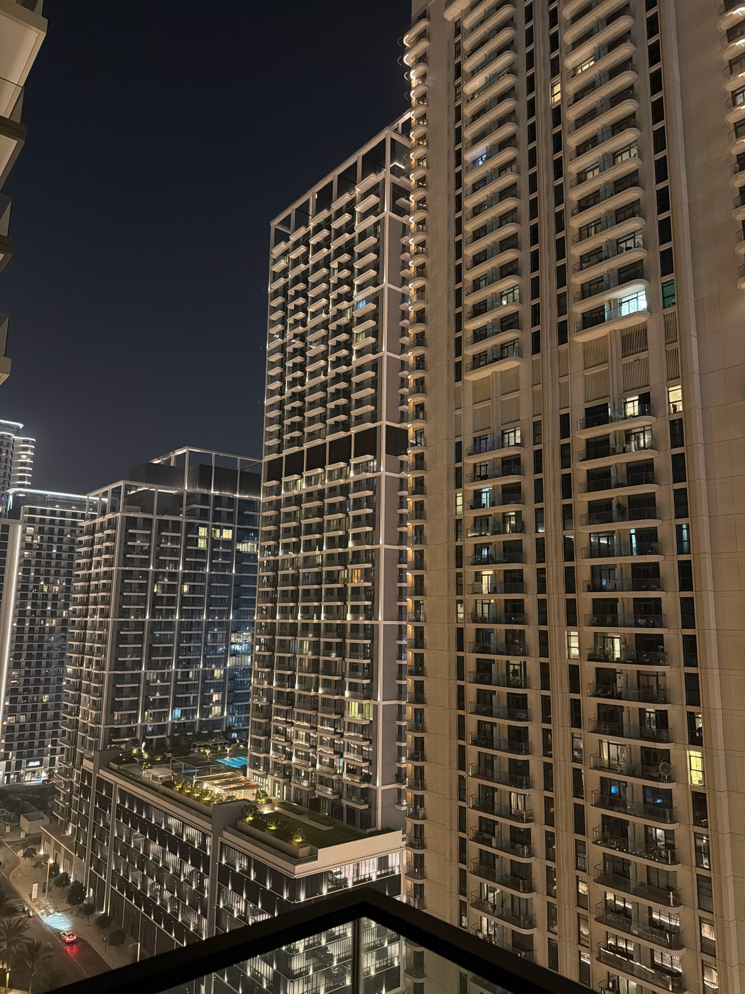 The image shows a night view of several modern high-rise buildings in Dubai, United Arab Emirates. The foreground features a close-up view of the facade of a tall, light-colored residential building, characterized by numerous balconies with white railings and dark glass windows. The building is illuminated, with lights visible in many of the windows, suggesting occupancy. To the left and in the background, other tall buildings are visible, some with intricate, grid-like architectural designs and illuminated windows and exterior lighting. One building in the mid-ground has a distinctive rooftop area with greenery and a swimming pool, also illuminated. The sky above is dark, indicating it is nighttime. At the bottom left of the image, a street scene is partially visible with a red car driving on a wet road, reflecting the streetlights. Palm trees can be seen in the lower left corner. The overall scene depicts a dense urban landscape with contemporary architecture, characteristic of Dubai's skyline.
