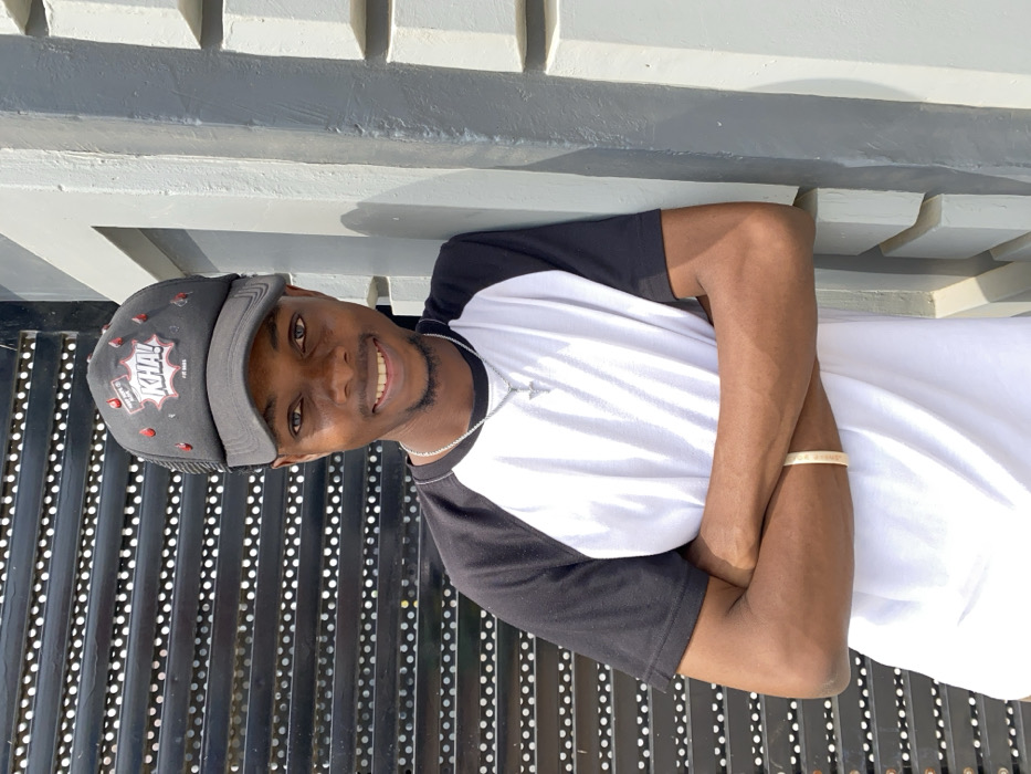 A young male subject is depicted outdoors in Joma, Ghana, facing the camera with a smile. He wears a black baseball cap featuring a "KHA!" speech bubble graphic and red drop-like embellishments, a black and white raglan-style t-shirt, a silver-colored chain with a cross pendant, and a light-colored bracelet on his right wrist. His arms are crossed over his chest. The background on the left consists of a dark, horizontally slatted structure. On the right, the subject leans against a light grey concrete wall with horizontal protrusions. The scene is illuminated by direct, bright natural light.