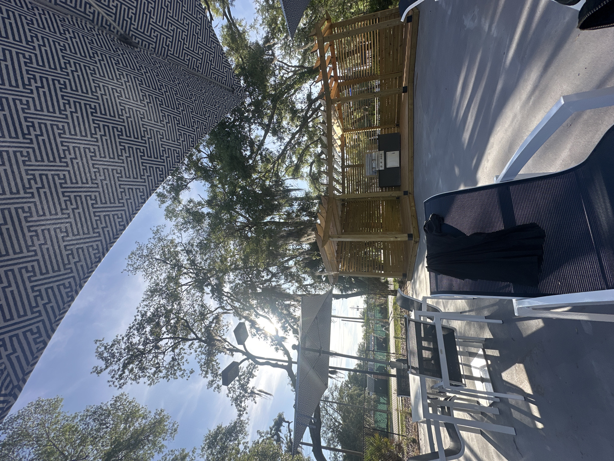 The image shows an outdoor patio area, likely in a residential or recreational setting. The location is in the city of University, United States of America. There is a partial view of a large patio umbrella with a geometric pattern in the foreground. Beyond the umbrella, there is a clear view of a wooden pergola structure containing an outdoor kitchen area with a grill. A set of outdoor dining chairs and a chaise lounge are visible on the right side of the image. The chaise lounge has a black fabric draped over it. The ground is concrete and appears to be wet in places. Large trees with Spanish moss are visible in the background, suggesting a southern climate. The sky is blue with some clouds. The scene is well-lit, and it appears to be a sunny day.