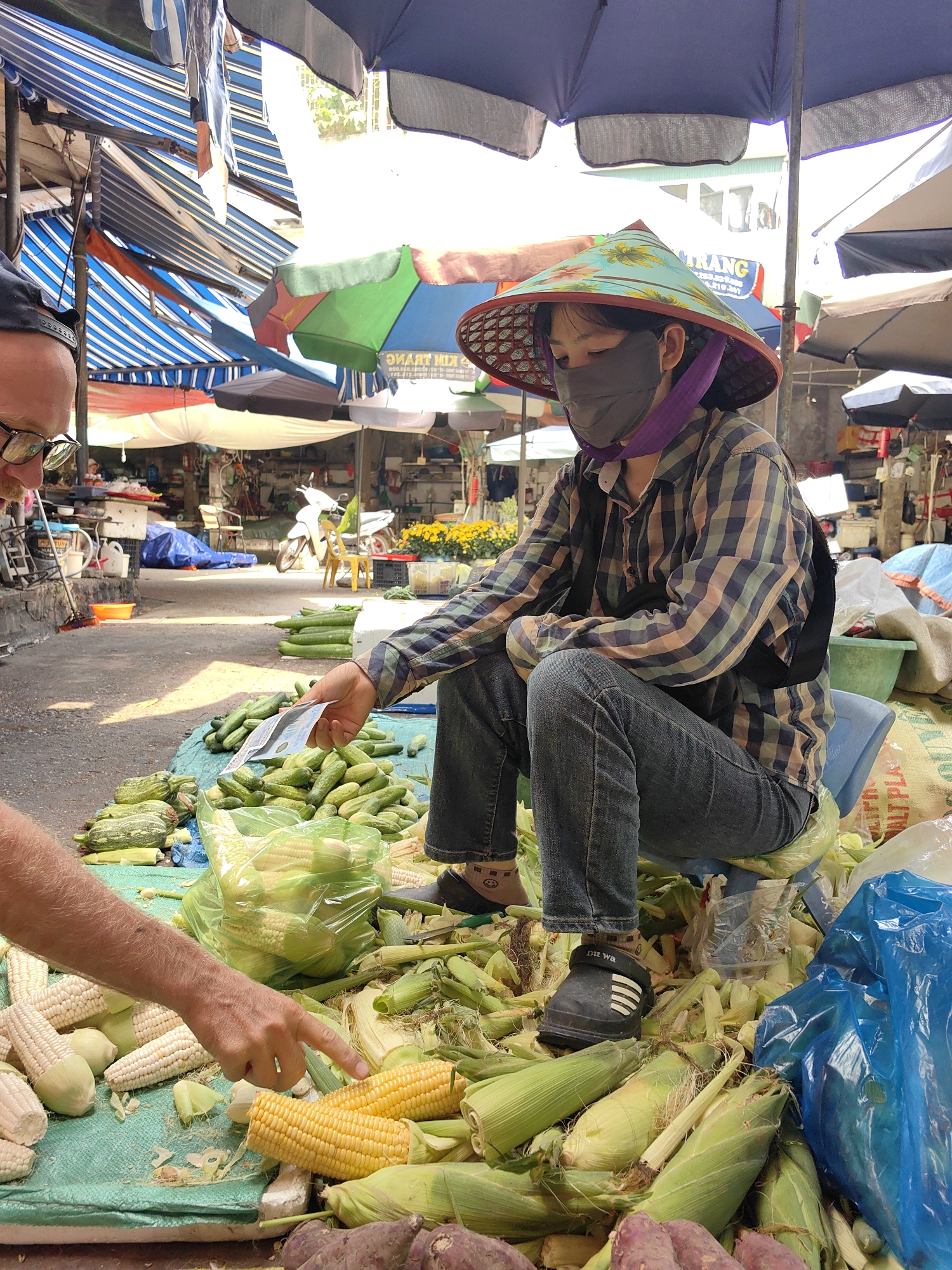 The image shows a woman in a traditional conical hat and face mask sitting at a market stall in Ha Long Ward, Vietnam. She is wearing a plaid shirt and jeans and is seated on a small blue plastic stool, surrounded by produce. The vendor is holding out money while an approaching customer gestures with his finger to several cobs of corn. They are surrounded by sweet potatoes, cucumbers, and corn in various states (husked, unhusked, and bagged). Umbrellas and awnings provide shade overhead.