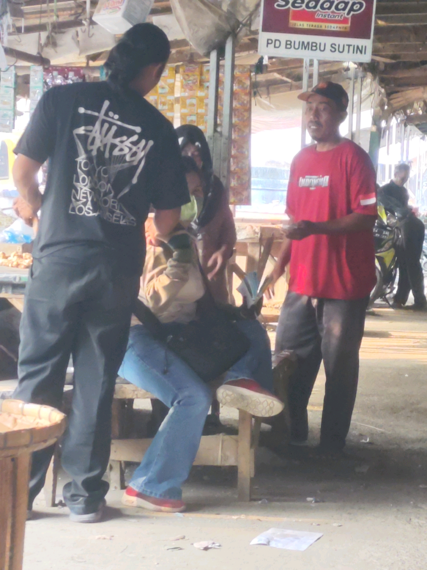 The image depicts a street market scene in Karawang, Indonesia. In the foreground, a person wearing a black t-shirt with a white graphic print and black pants stands to the left. To their right, a person in blue jeans and red-soled shoes is seated on a wooden bench. Another individual in a red t-shirt and dark pants stands to the right, facing the seated person and holding money. Behind them, a motorcycle is visible, and further in the background, stalls and goods are displayed under a makeshift roof. A sign with text that appears to be "SEDAP INSTANT" and "PD BUMBU SUTINI" is also visible overhead. The environment suggests a bustling, outdoor market setting.