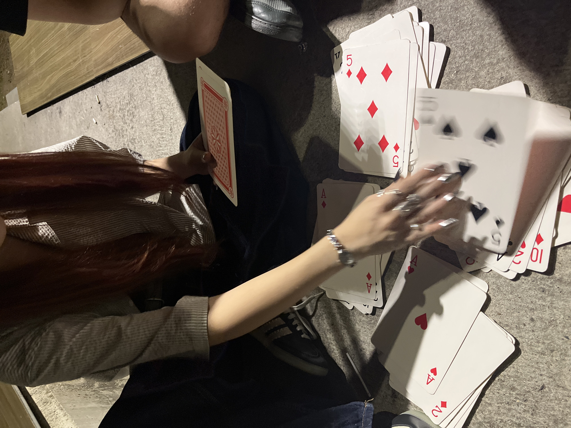 A person with long, reddish-brown hair, wearing a striped shirt and dark pants, is sitting on a textured surface, possibly concrete or asphalt, surrounded by scattered playing cards. They are holding a deck of cards in their left hand and their right hand is reaching towards another pile of cards. Several cards are visible, including the Ace of Diamonds, a 2 of Diamonds, a 4 of Diamonds, and a 5 of Diamonds. The environment appears to be outdoors, possibly in a park or public space, with a wooden bench visible in the background. The lighting suggests it is either dusk or nighttime. There is no identifiable information in the image that places the scene in Vratsa, Bulgaria.
