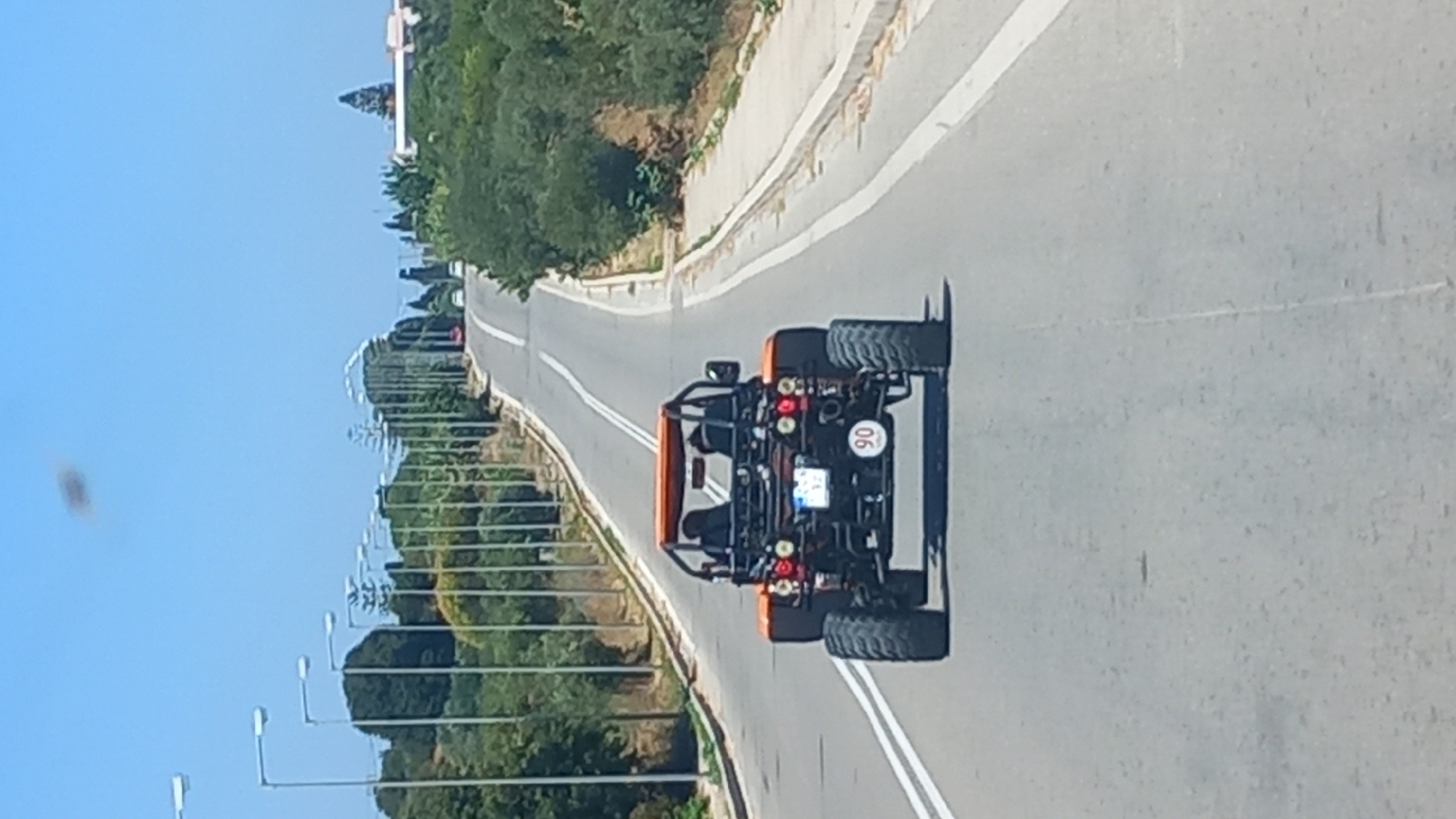 A low-angle, vertical, medium shot captures the rear view of an orange off-road buggy driving on a paved road. The buggy has a roll cage, two seats occupied by unseen figures, and large tires. A circular speed limit sign with "90" is visible on the rear of the buggy. The road has white lane markings and is flanked by a row of streetlights on the left and a grassy embankment with trees and vegetation on the right. In the distance, more streetlights and trees line the winding road as it ascends a hill. The sky is clear and blue, with a dark, blurry object present in the upper left quadrant of the frame.