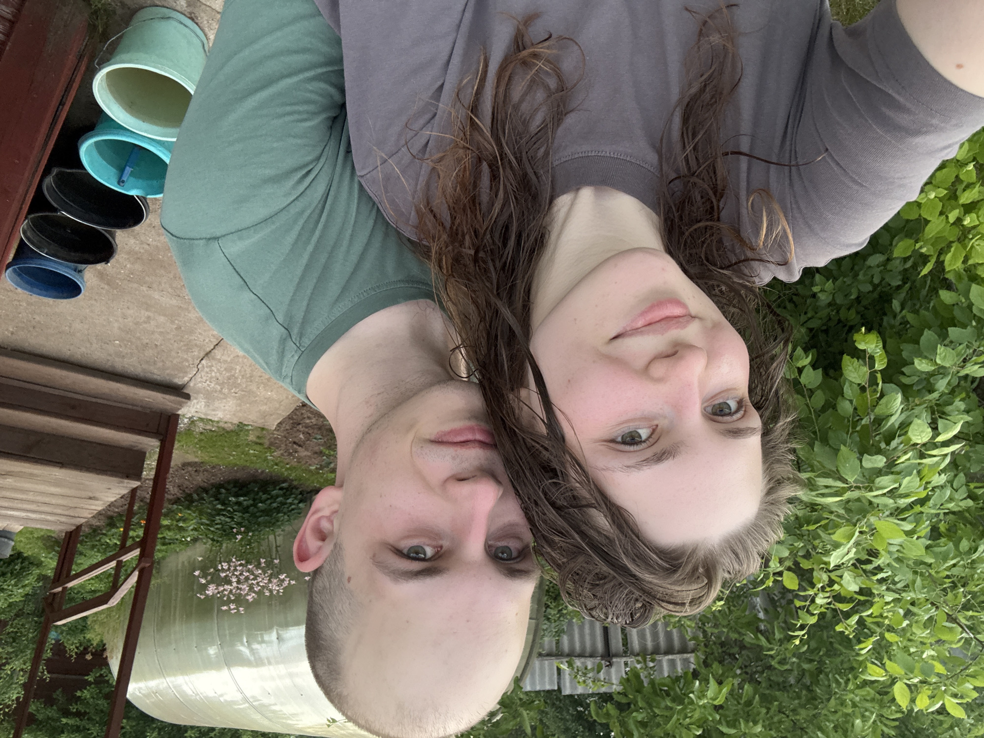 In the city of Дресвяново, Russia, a young couple is taking a selfie outdoors. The woman, with her hair pulled up, is in the foreground, facing the camera with a slight smile. Her skin appears flushed, and she has light-colored eyes. To her right, and slightly behind her, is a man with a shaved head and light-colored eyes, also looking towards the camera. They are both wearing casual t-shirts, the woman in grey and the man in green. The background reveals a lush green garden with thick foliage. To the right, a rustic wooden staircase leads up to a structure, possibly a porch. Further in the background, a translucent greenhouse stands prominently, with a cluster of pink flowers blooming in front of it. A row of buckets, varying in color from blue to green and black, are lined up near the staircase, some filled with water. The overall scene suggests a relaxed, domestic setting in a rural or suburban environment.