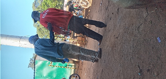 The image shows two men standing in a dusty, open-air environment. The man on the left is wearing a blue shirt, khaki pants, and a blue hat. The man on the right is wearing a red sweatshirt with the word "KENYA" written on the back, black pants, and a black hat. They appear to be having a conversation and are gesturing with their hands. There is a tall, white pole in the background. The environment appears to be a casual outdoor setting. The coordinates, 0.562 latitude and 34.805 longitude, place the location in the town of Kivaywa, Kenya.