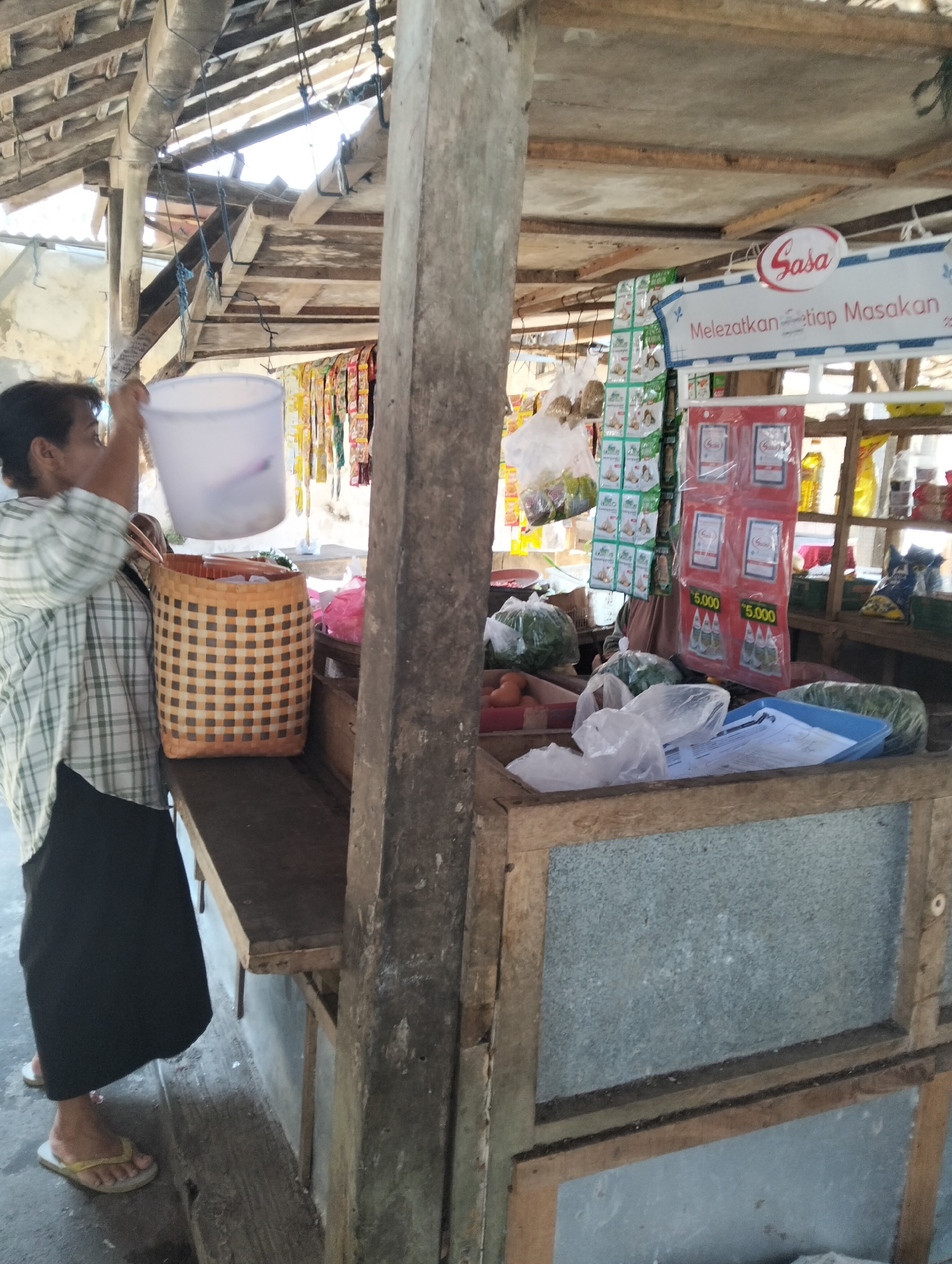 An outdoor market stall in Muncar, Indonesia, shows a woman in a plaid shirt and black skirt holding a white plastic bucket over a woven basket. Various goods, including bagged spices, vegetables in plastic, and bottled items, are displayed on shelves and counters. A sign with the logo "Sasa" is visible. The stall has a rustic, wooden construction with a corrugated roof.