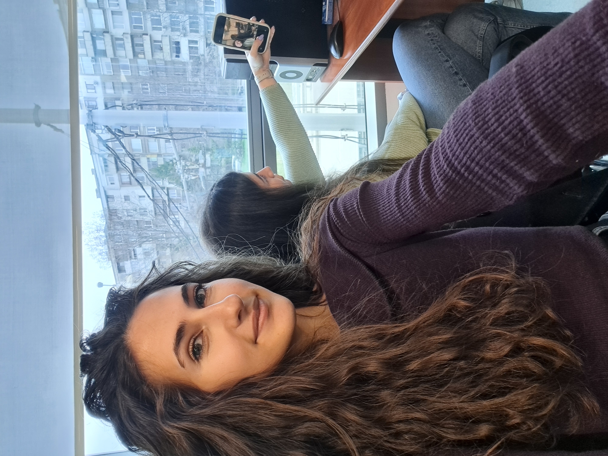 Two women are seated near a window. The woman in the foreground has long, curly brown hair, brown eyes, and a fair complexion. She is wearing a textured, dark purple top. The woman in the background, partially obscured, is taking a selfie with her mobile phone. She is wearing a light green, long-sleeved shirt. A computer monitor, mouse, and keyboard are visible on a desk next to them. Outside the window is an urban scene with buildings and power lines. The location is likely in Sofia, Bulgaria, given the style of architecture.