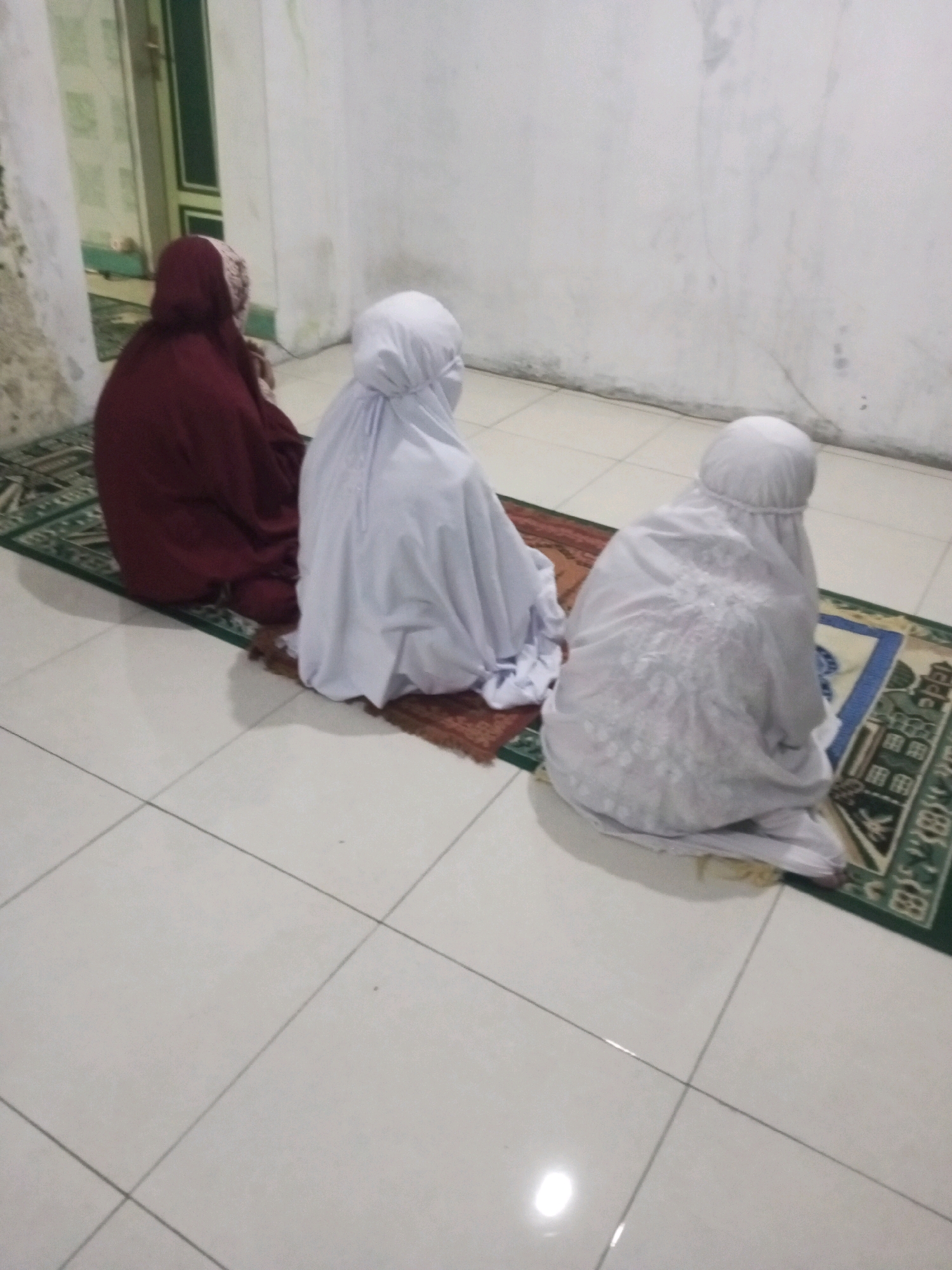 In Andongsili, Indonesia, three individuals are depicted performing Islamic prayer within what appears to be a residential setting. Two individuals are draped in white prayer garments, one with intricate white embroidery on the fabric, while a third individual is wearing a dark red garment. They are all seated on prayer rugs placed on a tiled floor. The room features white walls with some discoloration and a doorframe in the background. The overall lighting suggests an indoor environment, possibly at dusk or in a dimly lit room. The composition focuses on the act of prayer, with the individuals' backs to the viewer.
