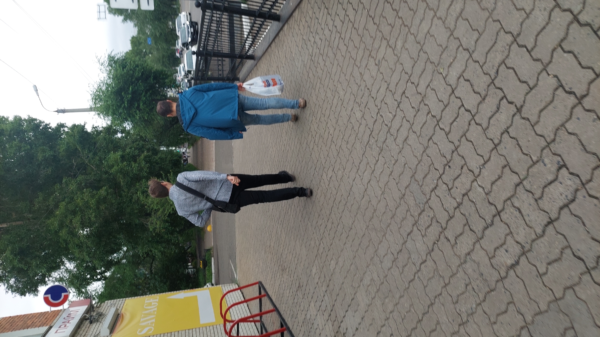 Two people are walking on a pedestrian sidewalk paved with interlocking concrete blocks. The person in the foreground is wearing a grey textured jacket and black pants, with a dark bag slung across their shoulder. The person behind them is wearing a blue jacket and blue jeans, carrying a white plastic shopping bag. Both individuals appear to be walking away from the camera.
The scene is outdoors, with lush green trees lining the left side of the sidewalk. A black metal fence runs along the right side of the sidewalk, separating it from what appears to be a road or parking area where several cars are visible. Overhead, the sky is overcast, suggesting a cloudy day.
On the left side of the image, partially visible, is a building with a sign. The sign has a blue and red circular logo with a white stripe and some red lines. Below this, there's a yellow sign with a white arrow pointing to the right, and the word "SAVAGE" written in white text. There is also text in what appears to be Cyrillic on the left side of the yellow sign. The overall impression is a typical urban or suburban street scene on a cool, cloudy day. No specific emotions are discernible from the subjects.