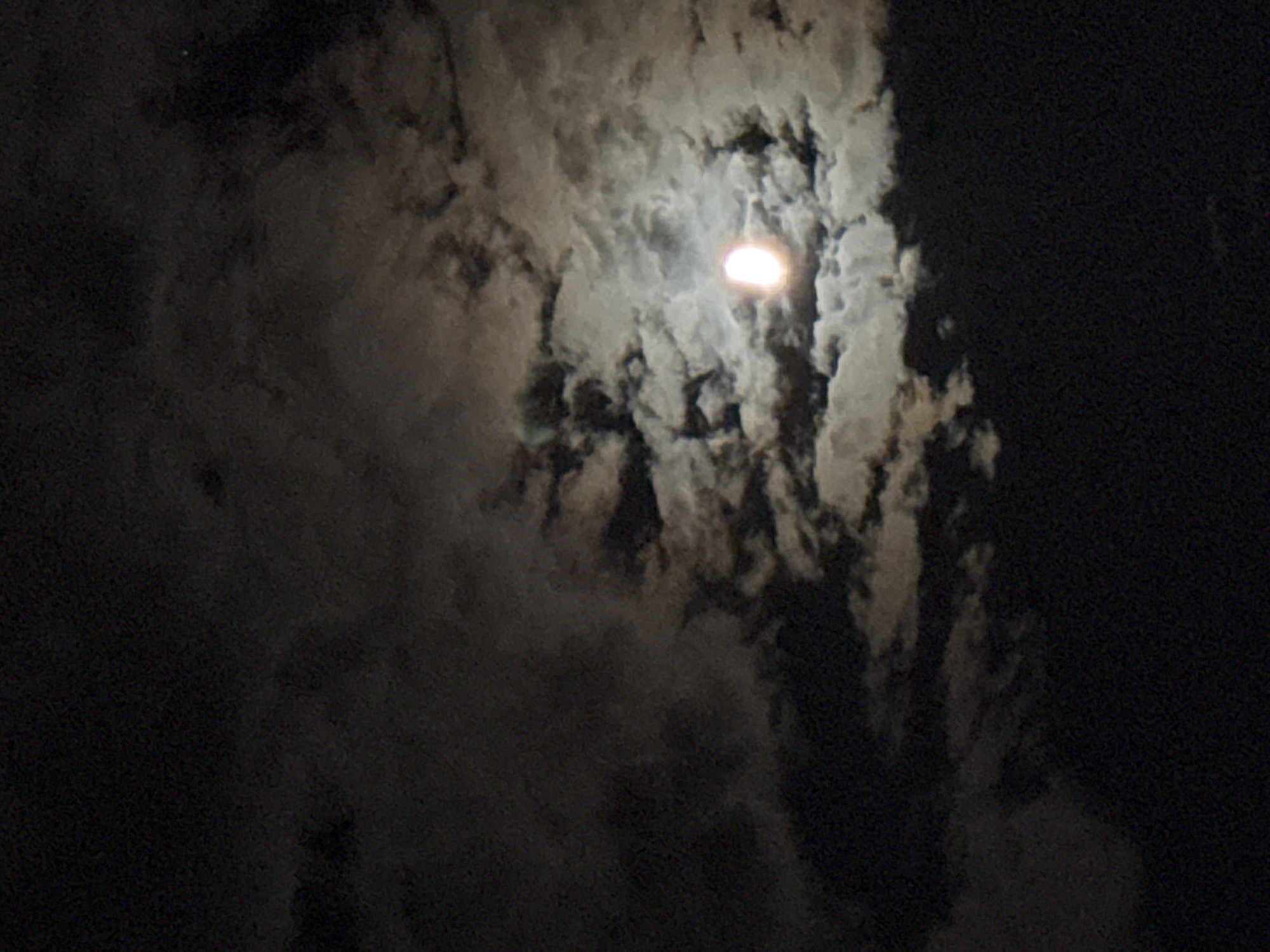 The image depicts a night sky over Rafailovići, Montenegro. A bright, semi-illuminated moon is visible, partially obscured by wispy clouds. The clouds vary in density, with some appearing dark and heavy, while others are lighter and translucent, allowing the moonlight to shine through. The overall impression is one of a slightly overcast night, with the moon's glow creating a soft, ethereal light. The lower portion of the image transitions to a dark, possibly terrestrial area, which is not clearly defined. There are no people or specific landmarks visible that would indicate a precise location within Rafailovići.