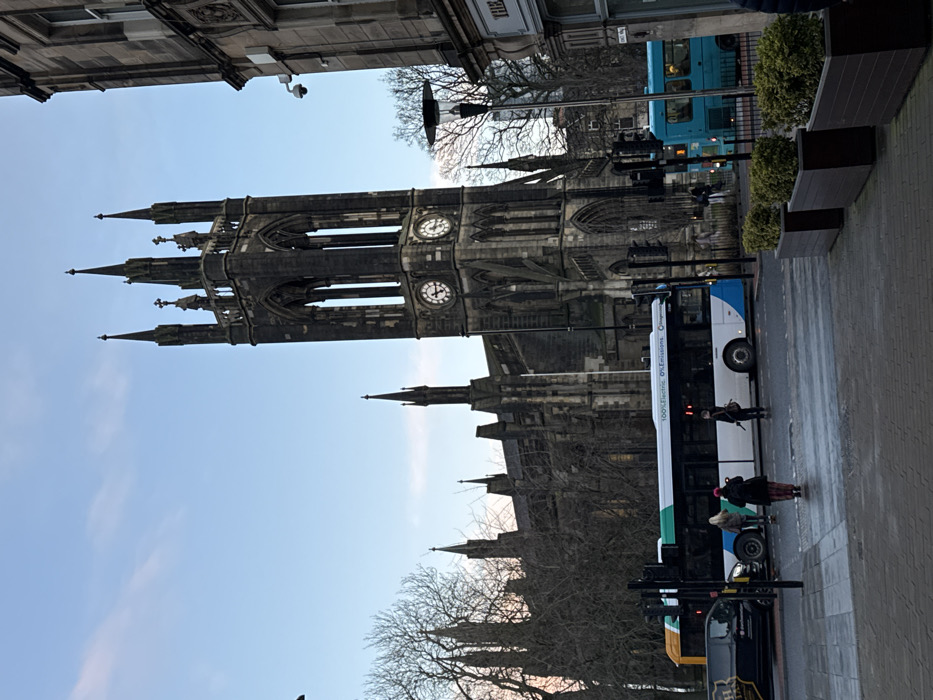 A tall, Gothic church with clock faces dominates the view. Trees stand bare in the foreground, partially obscuring lower portions of the church. A blue and white bus, along with other street vehicles, occupies the street level. Several pedestrians are visible near the bus. The sky is clear and light blue. Buildings border the scene at the top.