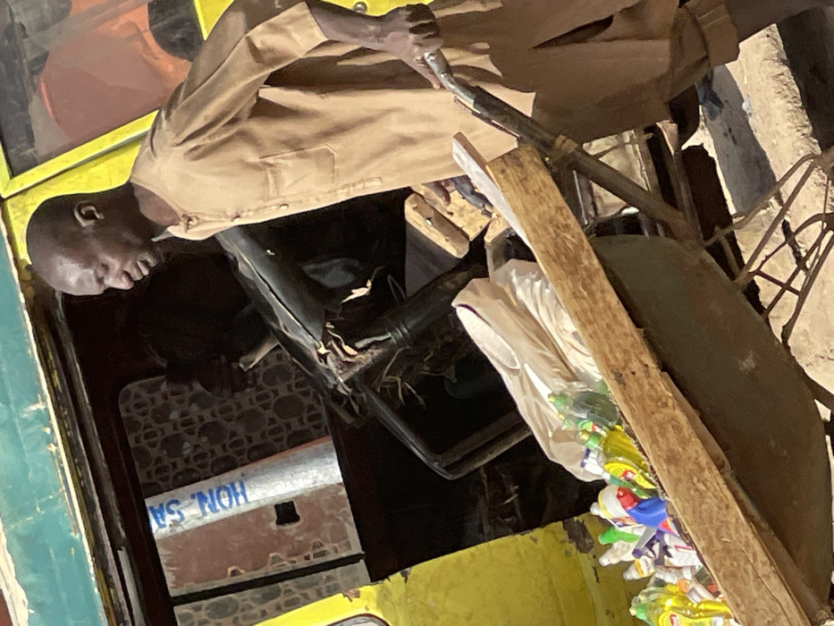 A bald male, dressed in a light brown tunic, pushes a metal wheelbarrow laden with numerous plastic bottles containing various colored liquids and white plastic bags. His right hand grasps the wheelbarrow handle as he moves leftward. In the background, a yellow and green vehicle, likely a bus or large van, displays an open interior with a dark steering wheel, a damaged seat, and a reflective pole inscribed with "HON-SA" in blue text. An obscured individual is visible within the vehicle's interior. The ground is dusty and uneven. The scene is located in Zaria, Nigeria.