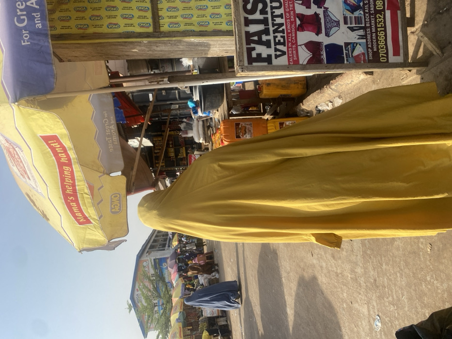 This image depicts a bustling outdoor market scene in Zaria, Nigeria, captured during bright daylight with clear skies, indicated by strong shadows and overhead light. The photograph is oriented vertically, rotated 90 degrees clockwise.
In the foreground, a person is largely obscured, draped in a vibrant yellow garment that covers most of their body. Their head is visible, also covered in yellow fabric. To the left, a large yellow market umbrella is prominent, displaying text such as "Mama's helping hand!" and branding for "ONGA." A blue umbrella is partially visible next to it. These umbrellas provide shade over market stalls.
The background reveals more of the market environment. To the right, a white signboard for "FAISAL VENTURES" advertises "DEALERS IN ALL KINDS OF GYALI, SHIRTS, CAMISOL, SKIN TIET." The sign also provides a "SHOP ADDRESS: BLOCK A15, S MODERN MARKET, SABON" and contact numbers "07036661532, 08". Behind this sign, a wall is covered with repeated "ONGA" branding. Various yellow jerry cans or containers are visible on the ground near the stalls. Further into the distance, other market stalls, goods, and several other people (partially obscured) can be seen, contributing to the lively atmosphere. A distant building with a "MEGA" sign is also visible under the clear sky. The ground is a paved surface, showing some minor debris.