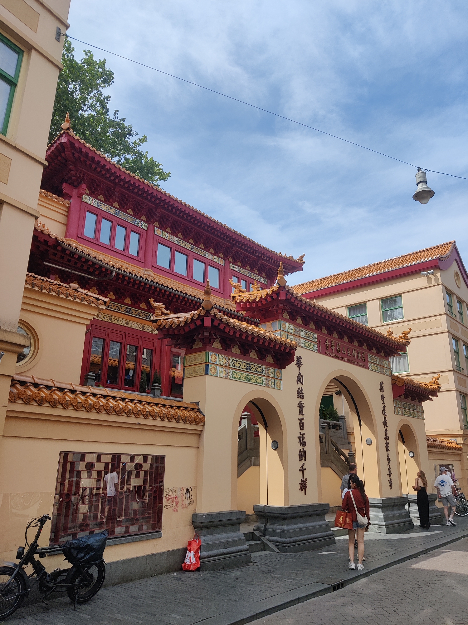 The image shows the Fo Guang Shan He Hua Temple in Amsterdam, Netherlands, on a sunny day. The temple has a traditional Chinese architectural style with red tiled roofs and ornate decorations. The main building is painted a light beige color. In the foreground, there is a street with cobblestone paving. To the left, a black bicycle with a black bag on the back is parked. A red garbage bag with a white logo is leaning against the wall to the right of the bicycle. Several people are visible walking in front of the temple's arched entrance. One woman in a red jacket and denim shorts is carrying a shopping bag. Further back, a man and woman are walking away from the camera. Another man in a white t-shirt and shorts is walking past a bicycle on the right side of the image. The sky is blue with some scattered white clouds, and a street lamp hangs from a wire above. The building also features Chinese characters inscribed above the arched entrance.