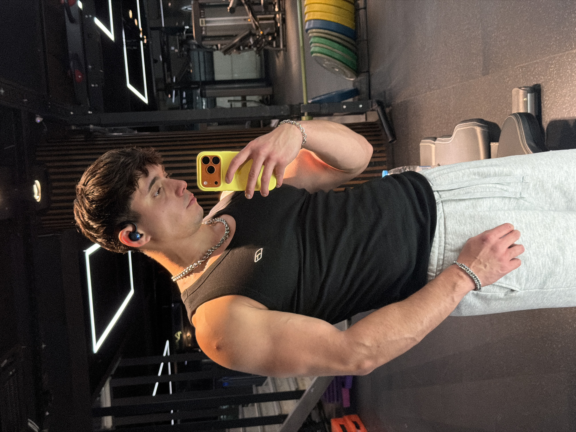 A young man in a black tank top and gray sweatpants stands in a gym taking a selfie with a yellow smartphone. He wears a silver chain necklace and matching bracelets on both wrists. He also has a blue earbud in his left ear. Gym equipment is visible in the background, including weight plates and workout machines. Modern geometric lighting fixtures adorn the walls. There is no information to determine the location in the city of Sofia, Bulgaria.