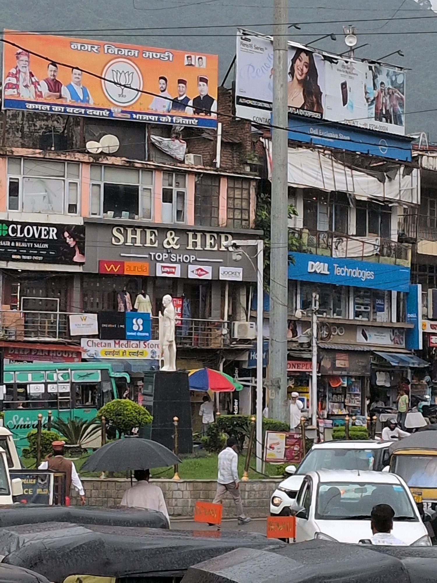 The image depicts a street scene in Mandi, India, likely a busy commercial area. A large banner at the top advertises the Nagar Nigam Mandi and features portraits of political figures, with the Bharatiya Janata Party (BJP) lotus symbol prominently displayed. Below the banner, a multi-story building houses various shops. On the left, a sign for "Clover Hair & Beauty Salon" is visible. Adjacent to it, a prominent sign reads "SHE & HER TOP SHOP," with smaller brand logos like "W," "Aurelia," and "Jockey" displayed below. Further to the right, a blue sign for "DELL Technologies" indicates an electronics store.
In the foreground, a statue of a person in a standing pose is positioned on a pedestal. The street is occupied by several vehicles, including auto-rickshaws and cars. People are seen walking and standing amidst the traffic. One man is holding a black umbrella, suggesting it might be raining or the sun is strong. The architecture appears to be a mix of traditional and modern elements, common in Indian cities. The overall impression is that of a lively urban environment with commercial activity.